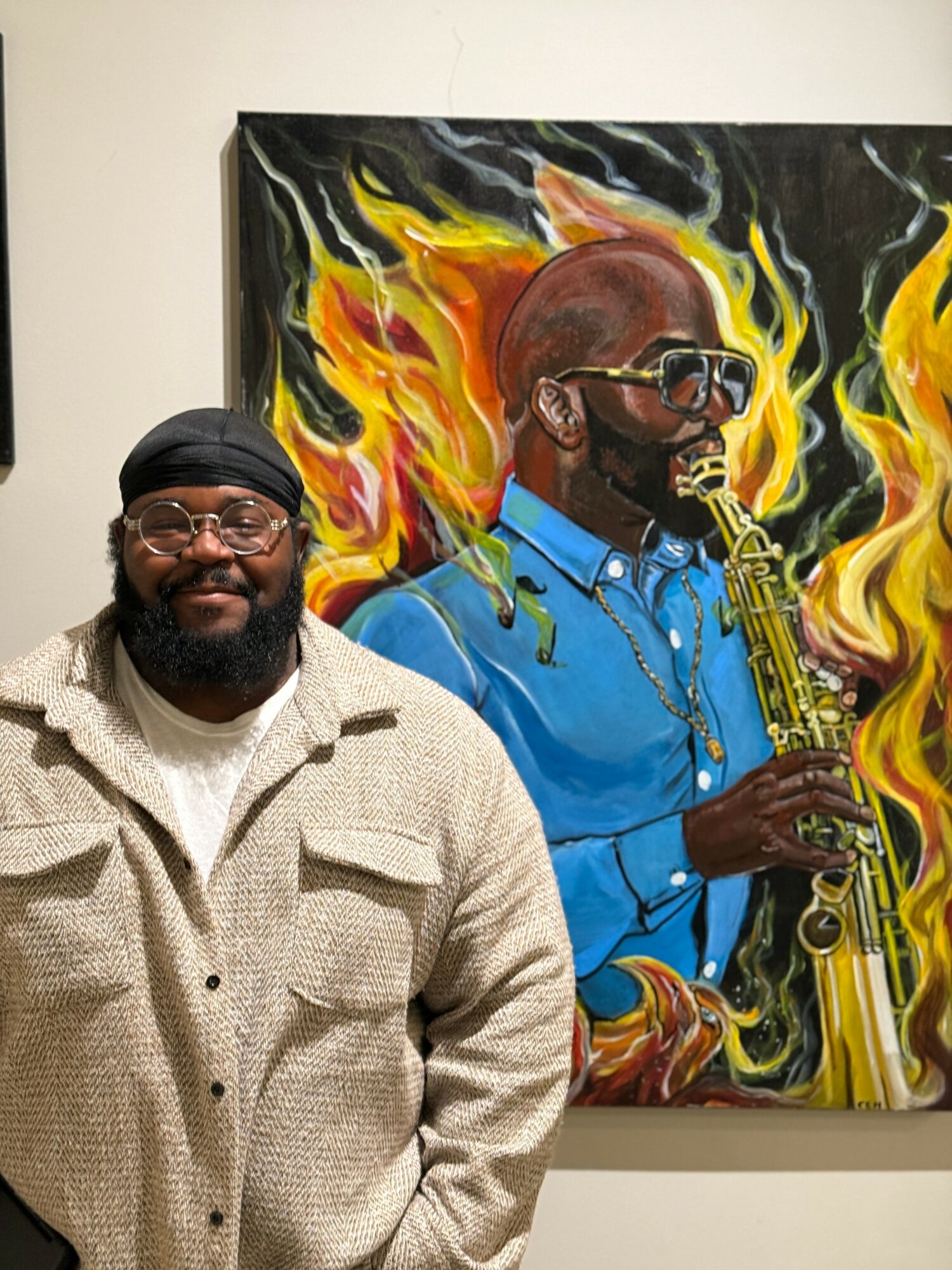 Man with glasses and a beard wearing a beige jacket standing in front of a colorful painting of a saxophonist with flames.