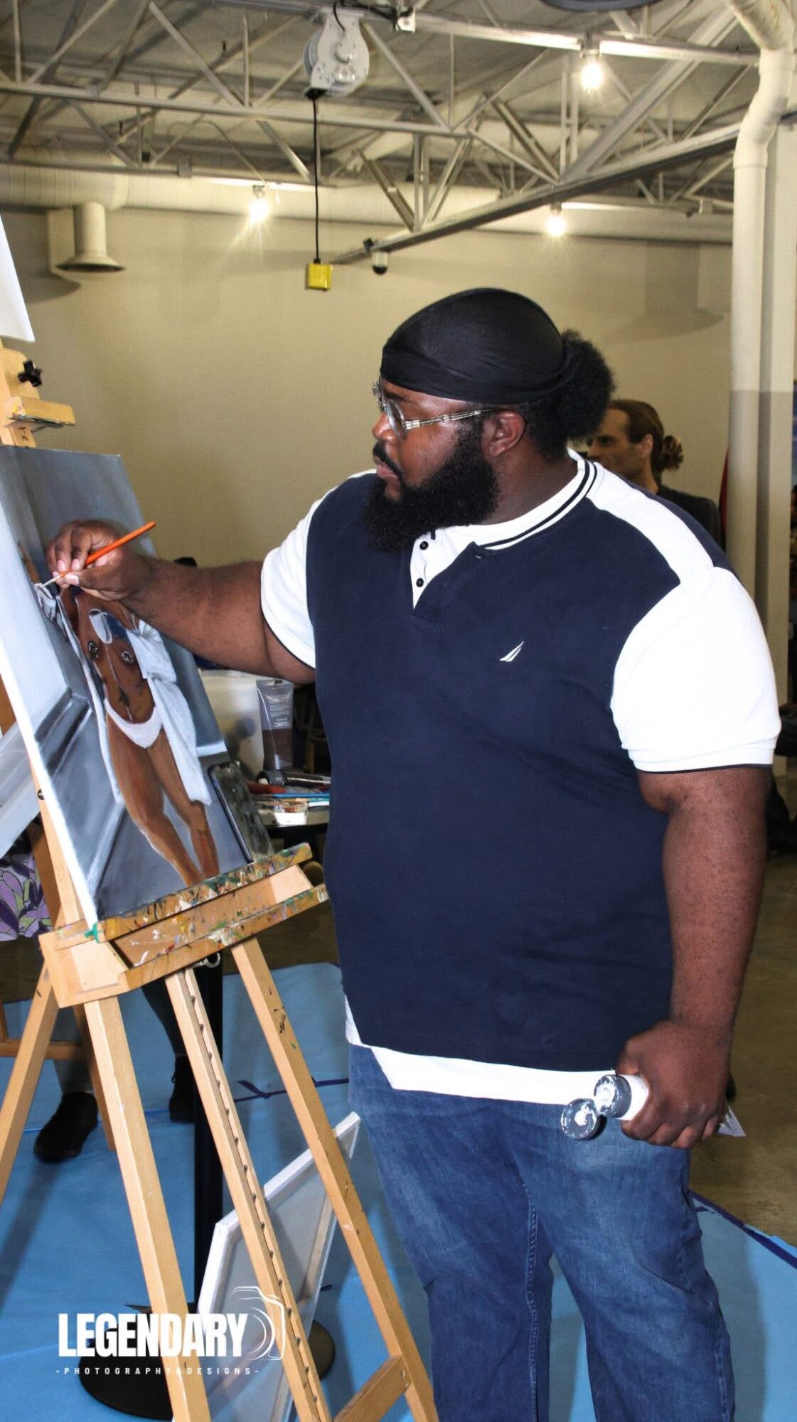 Person with glasses and a black head covering painting on a canvas at an indoor event.