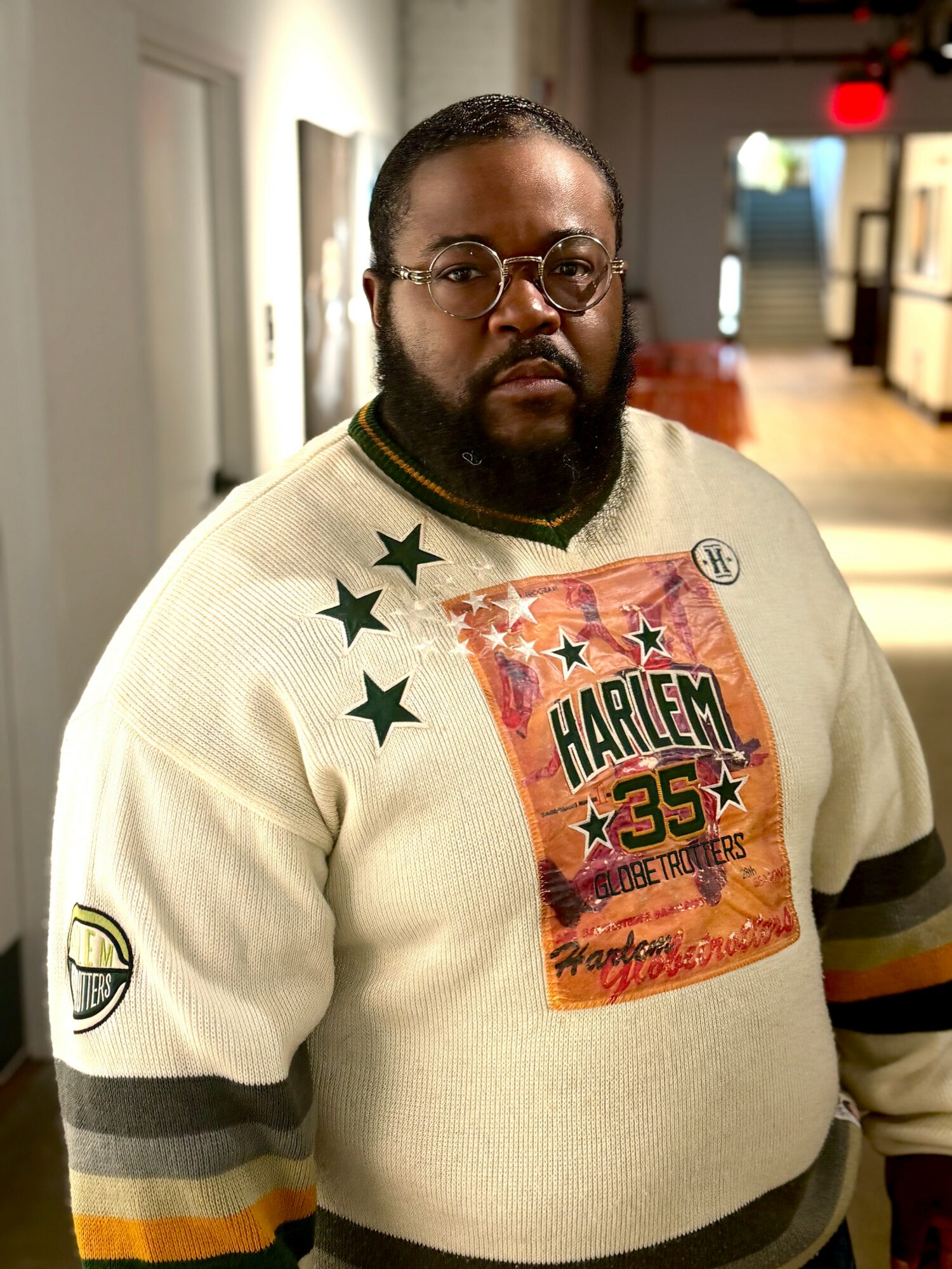 Man with glasses and beard wearing a cream sweater with a graphic design, standing indoors.