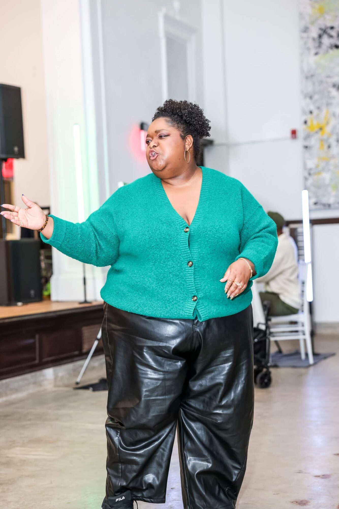 Woman with short curly hair wearing a teal cardigan and black leather pants, gesturing with her right hand in a room.
