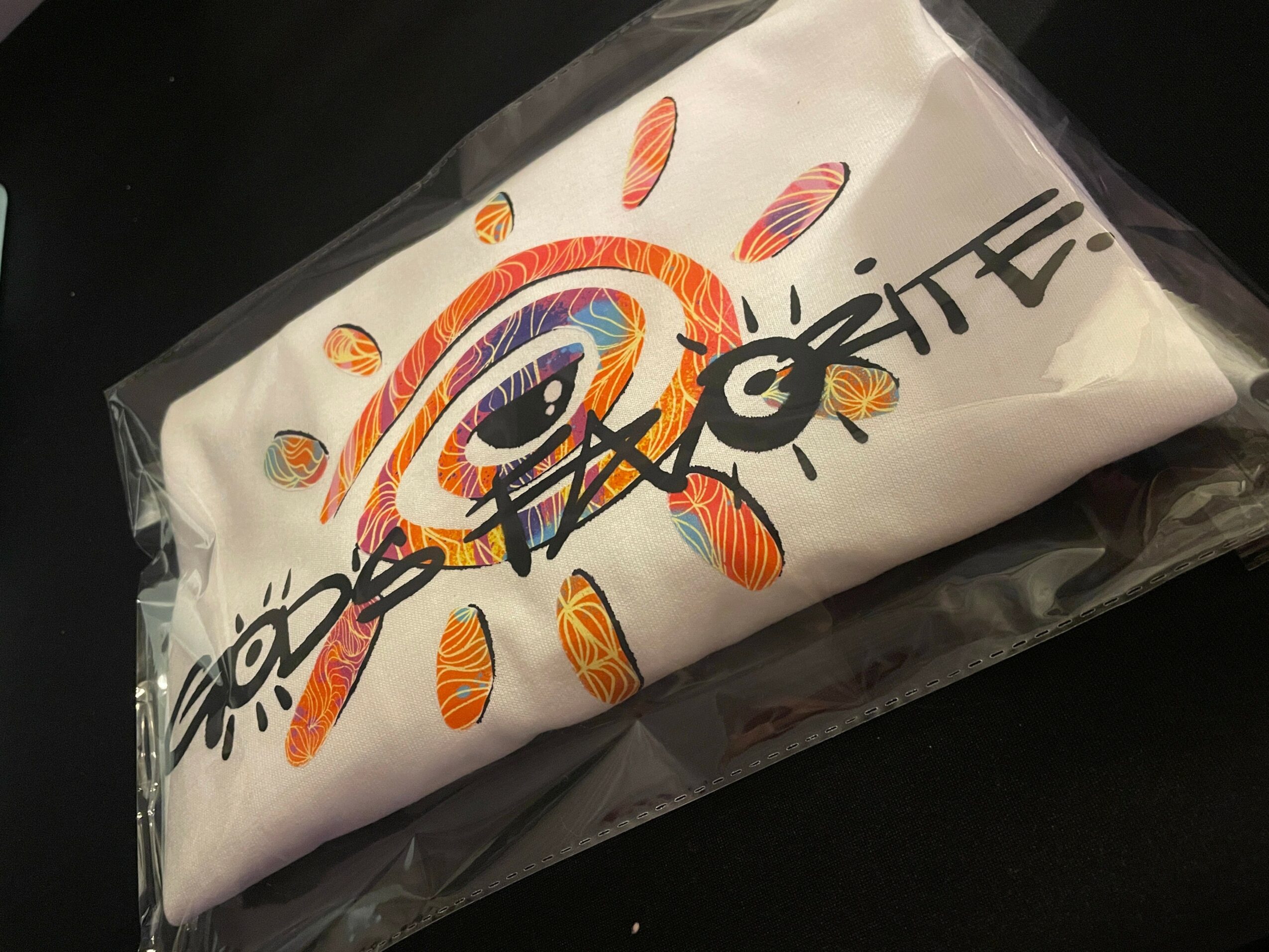 White fabric with colorful rainbow and sun design, partially covered by black text, inside clear plastic packaging.