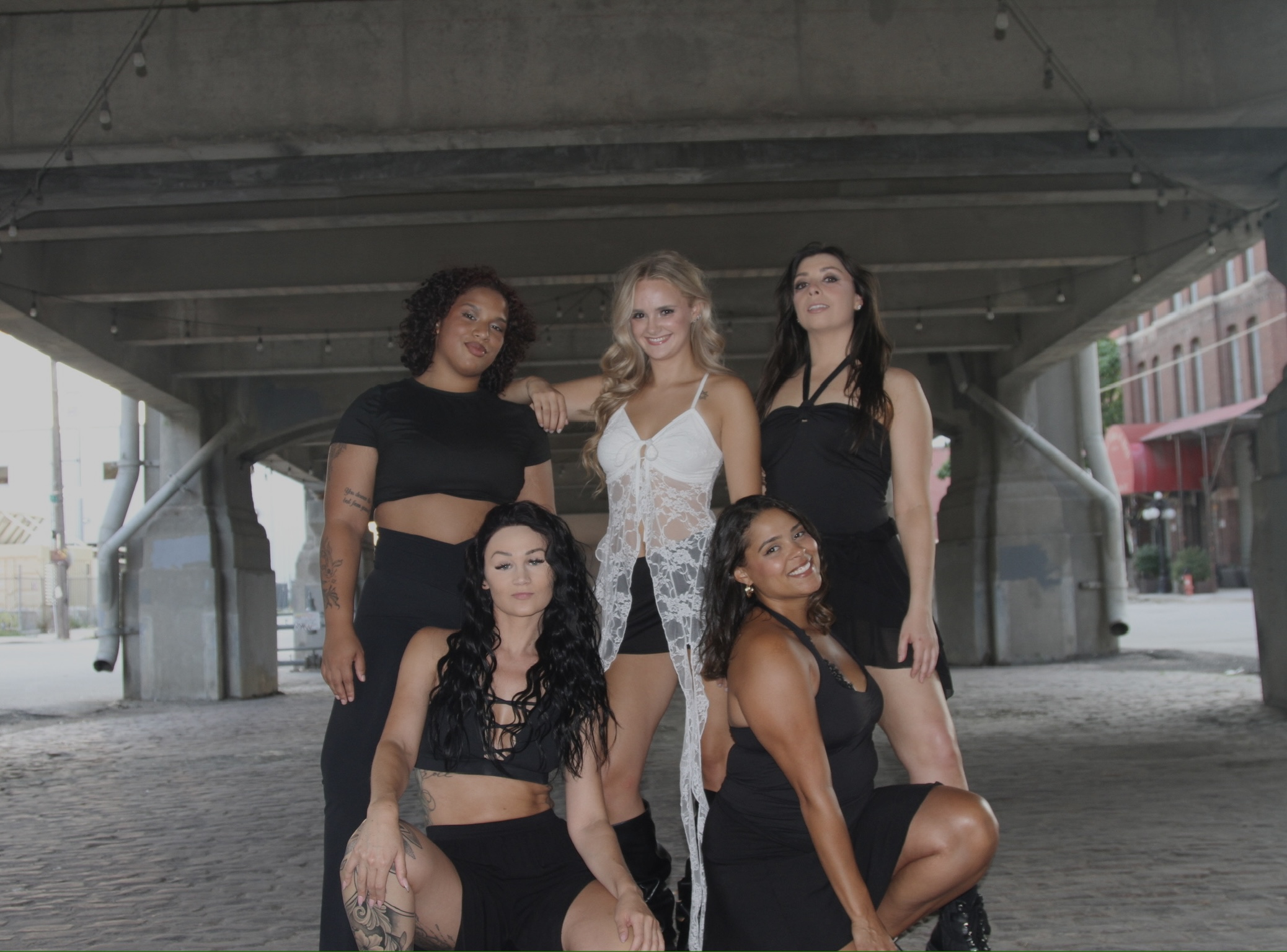 Five women posing under a bridge, some standing and some kneeling, in casual clothing.