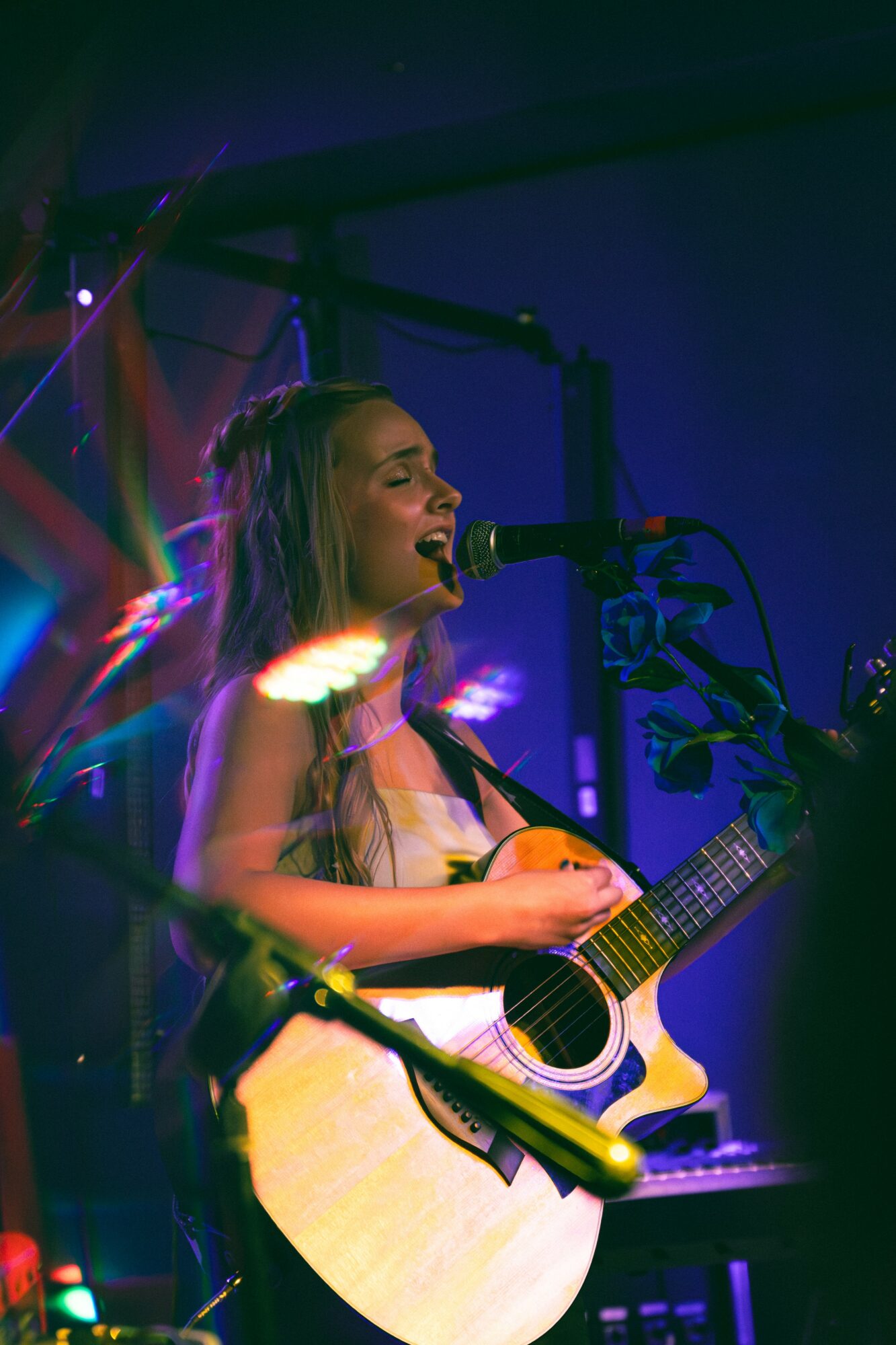 Young woman singing into a microphone while playing an acoustic guitar on stage with colorful lighting.