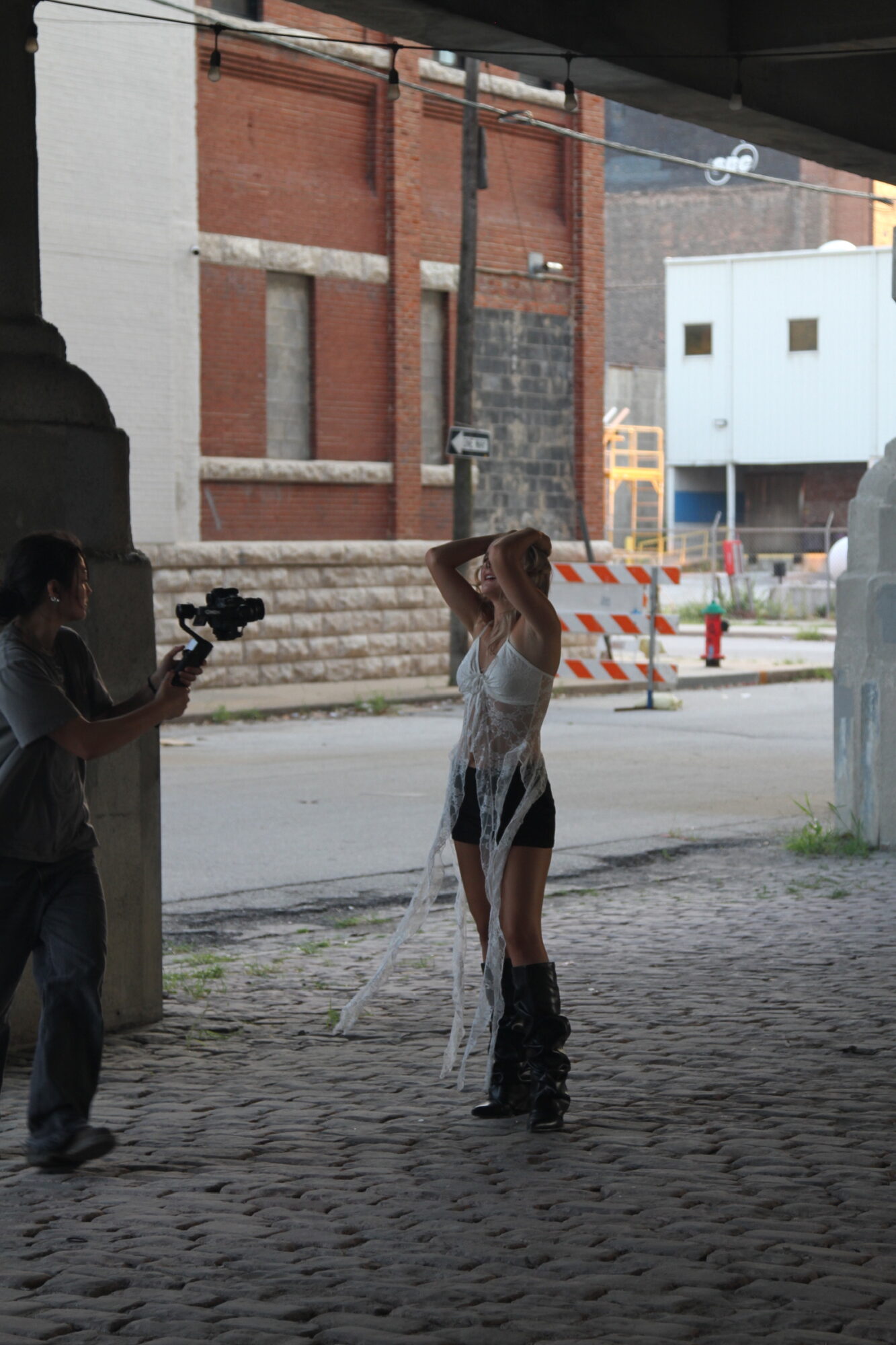 Person standing outdoors under a bridge, holding their head, with a person filming nearby, in an urban setting.