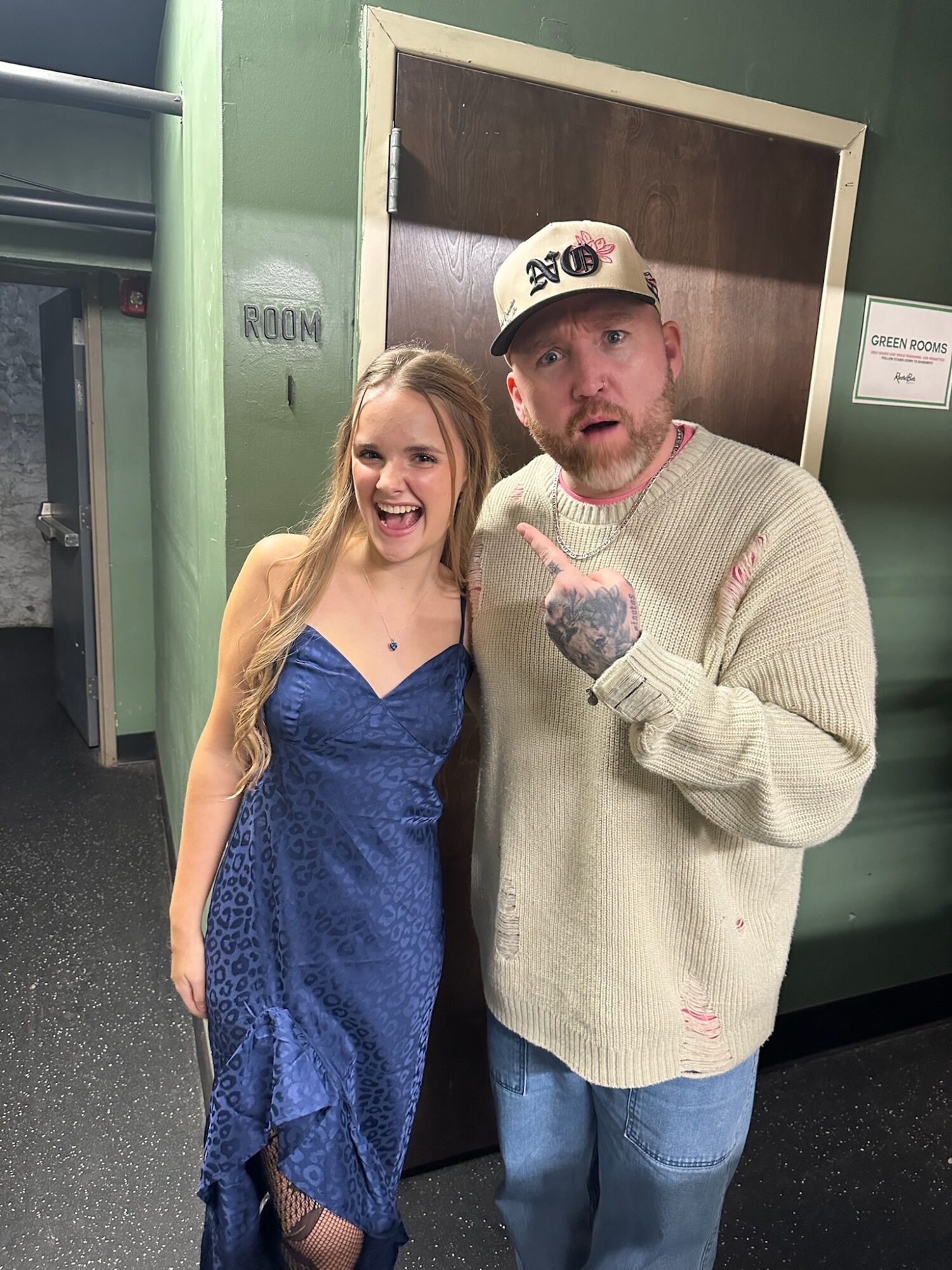 A smiling woman in a blue dress and a man in a beige sweater and cap pose together indoors.