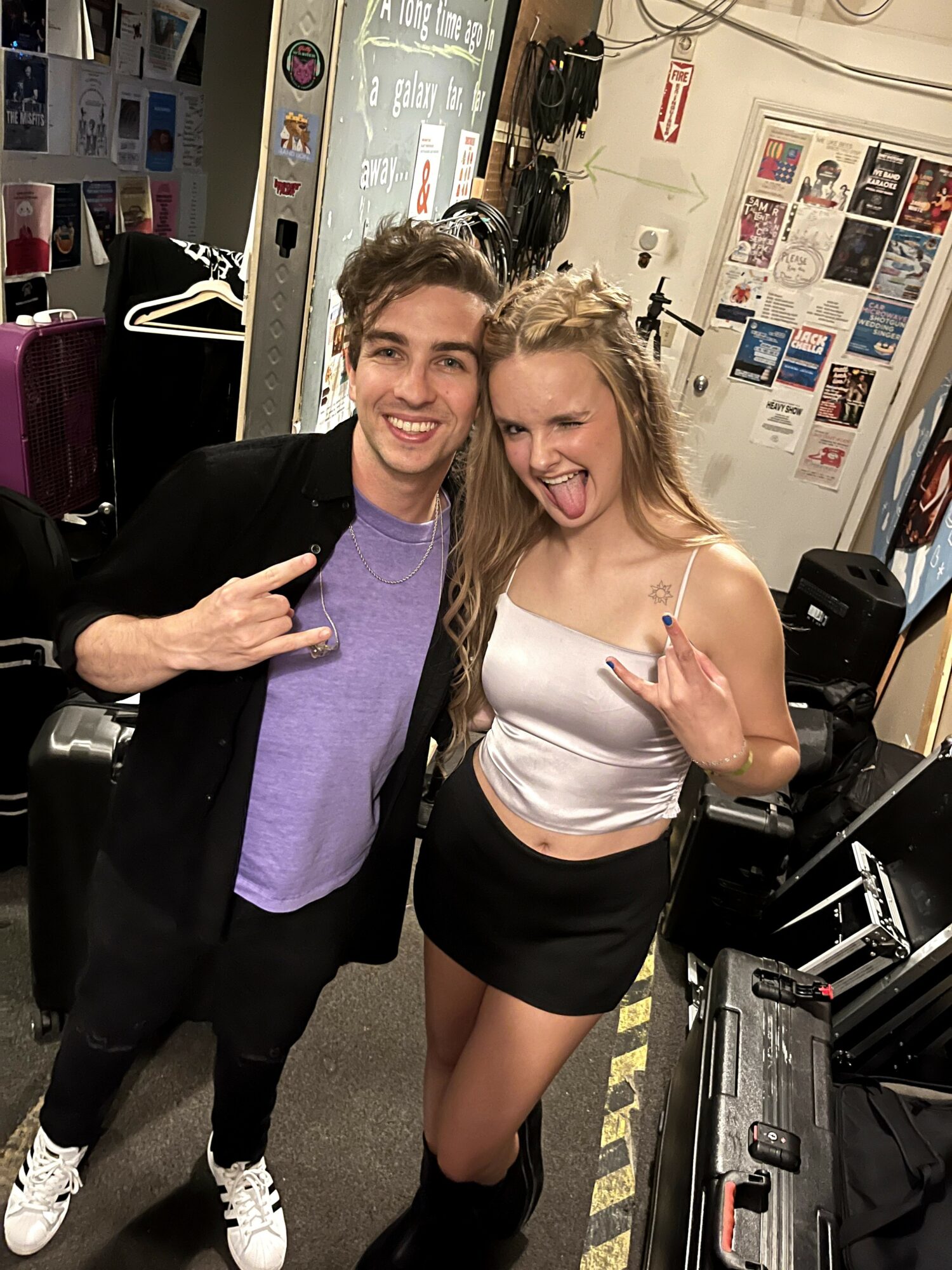 Two people smiling and making hand gestures in a room with posters and equipment, one wearing a black jacket and purple shirt, the other in a white top and black skirt.