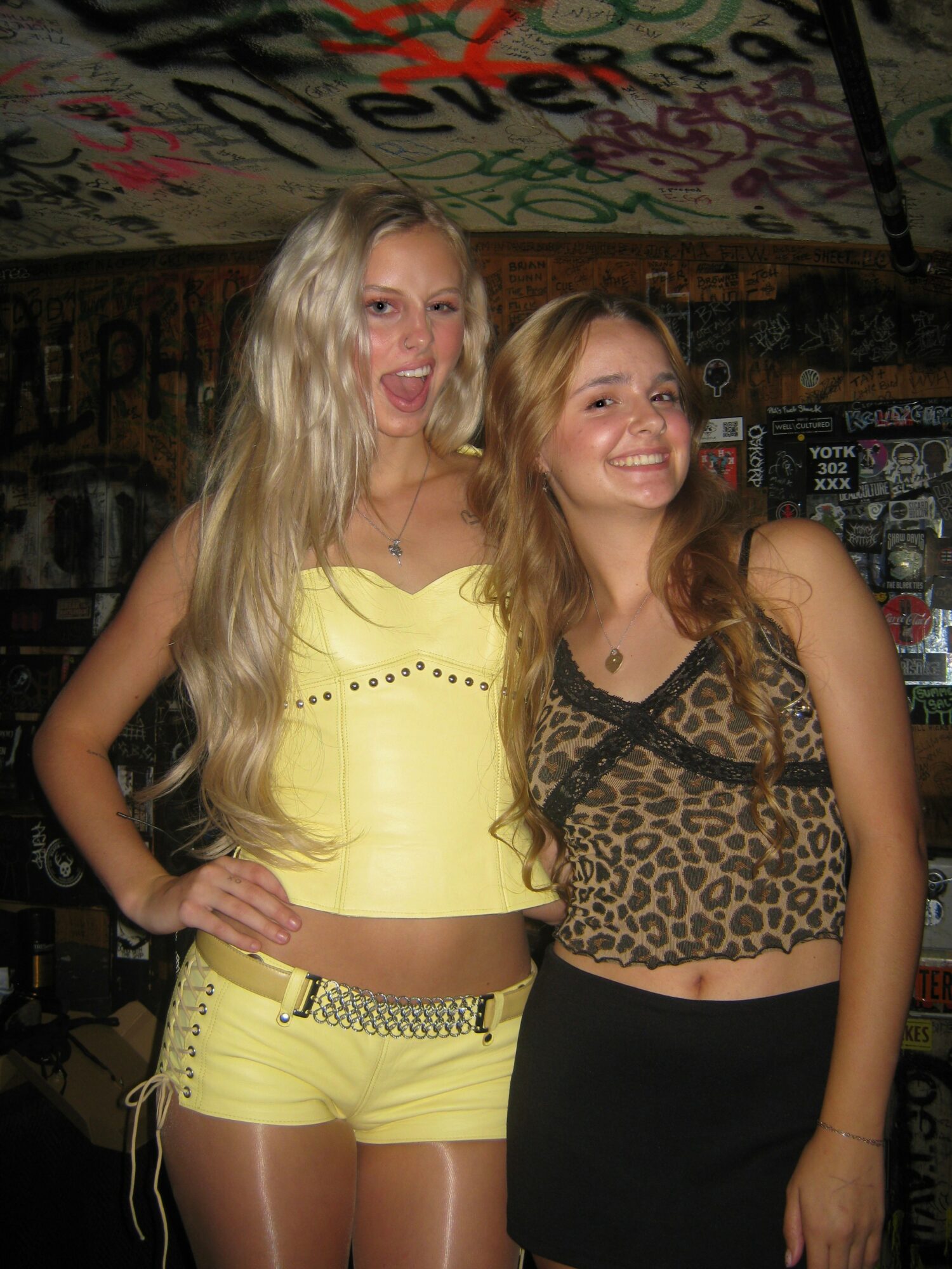 Two young women smiling, one in yellow shorts and top, the other in a leopard print top and black skirt, in a graffiti-covered setting.