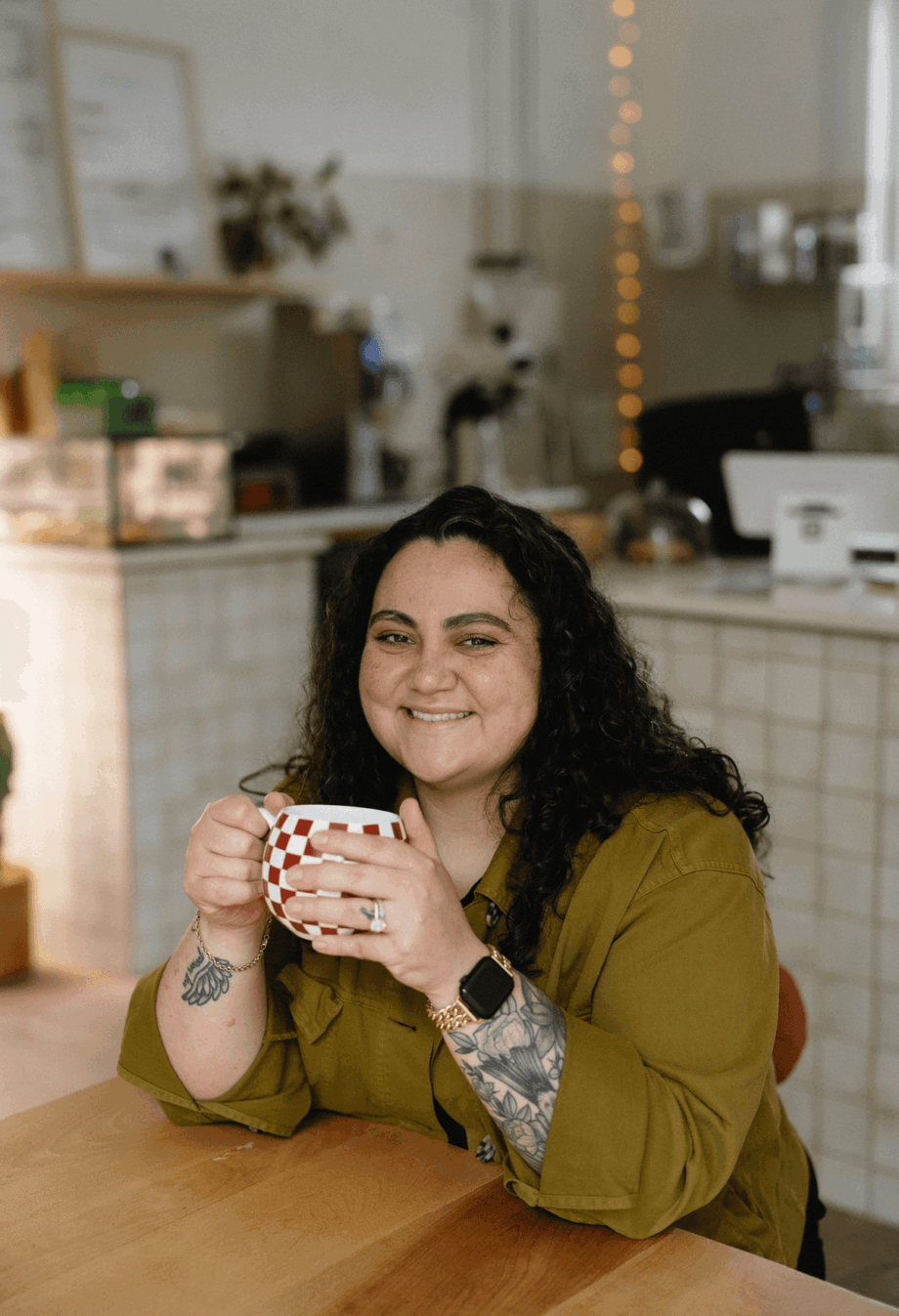 Woman with curly hair smiling, holding a mug, sitting at a table in a cafe, decorated with string lights.
