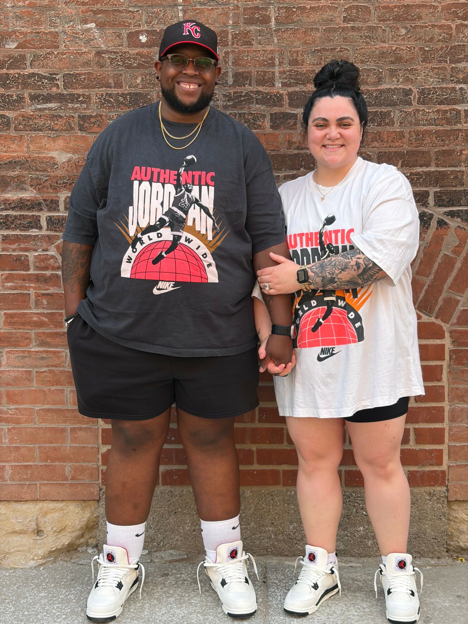 Two people stand side by side against a brick wall, smiling, wearing casual outfits and white sneakers.