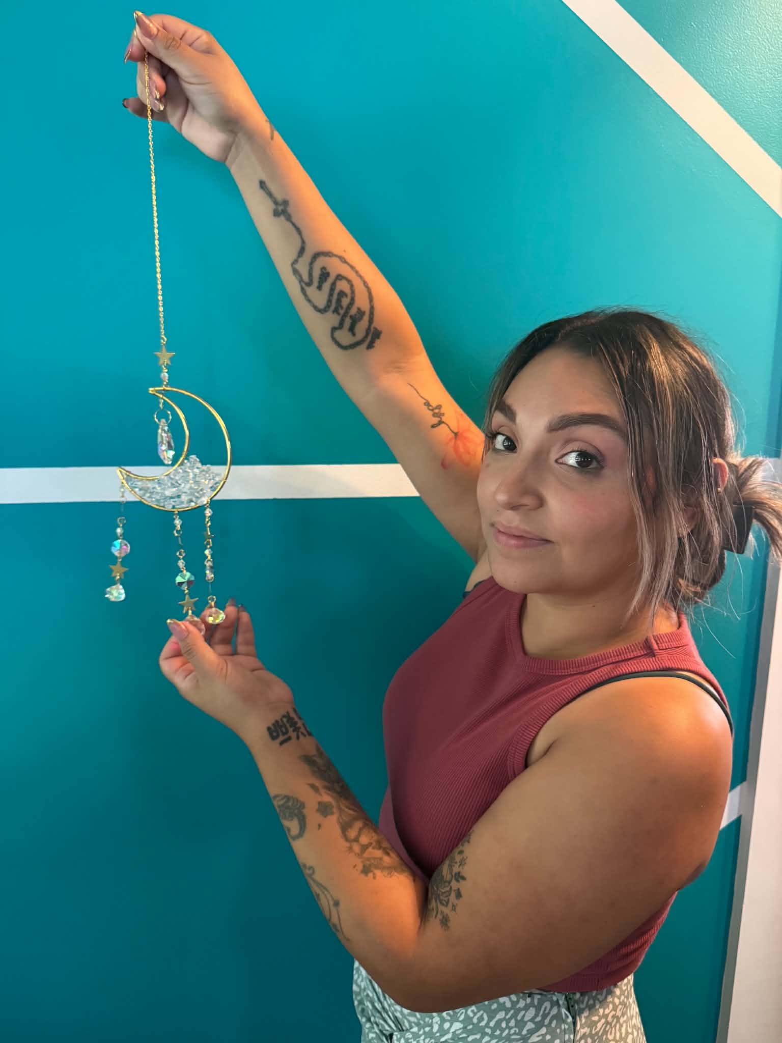 Young woman with tattoos holding a dreamcatcher decoration against a blue wall, smiling slightly, wearing a sleeveless top.