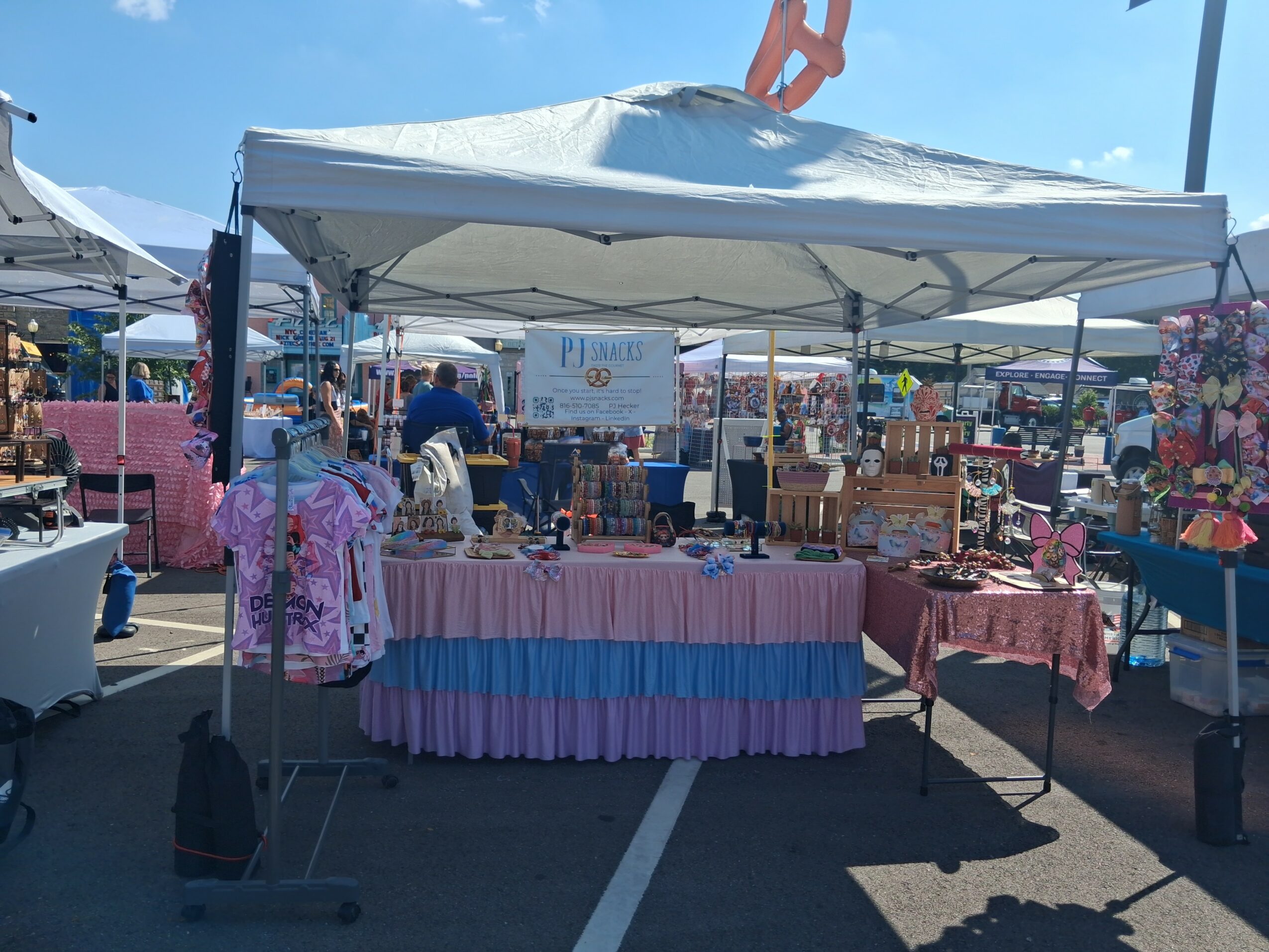 Outdoor market stall with a white canopy, displaying colorful clothing and accessories, under a blue sky.