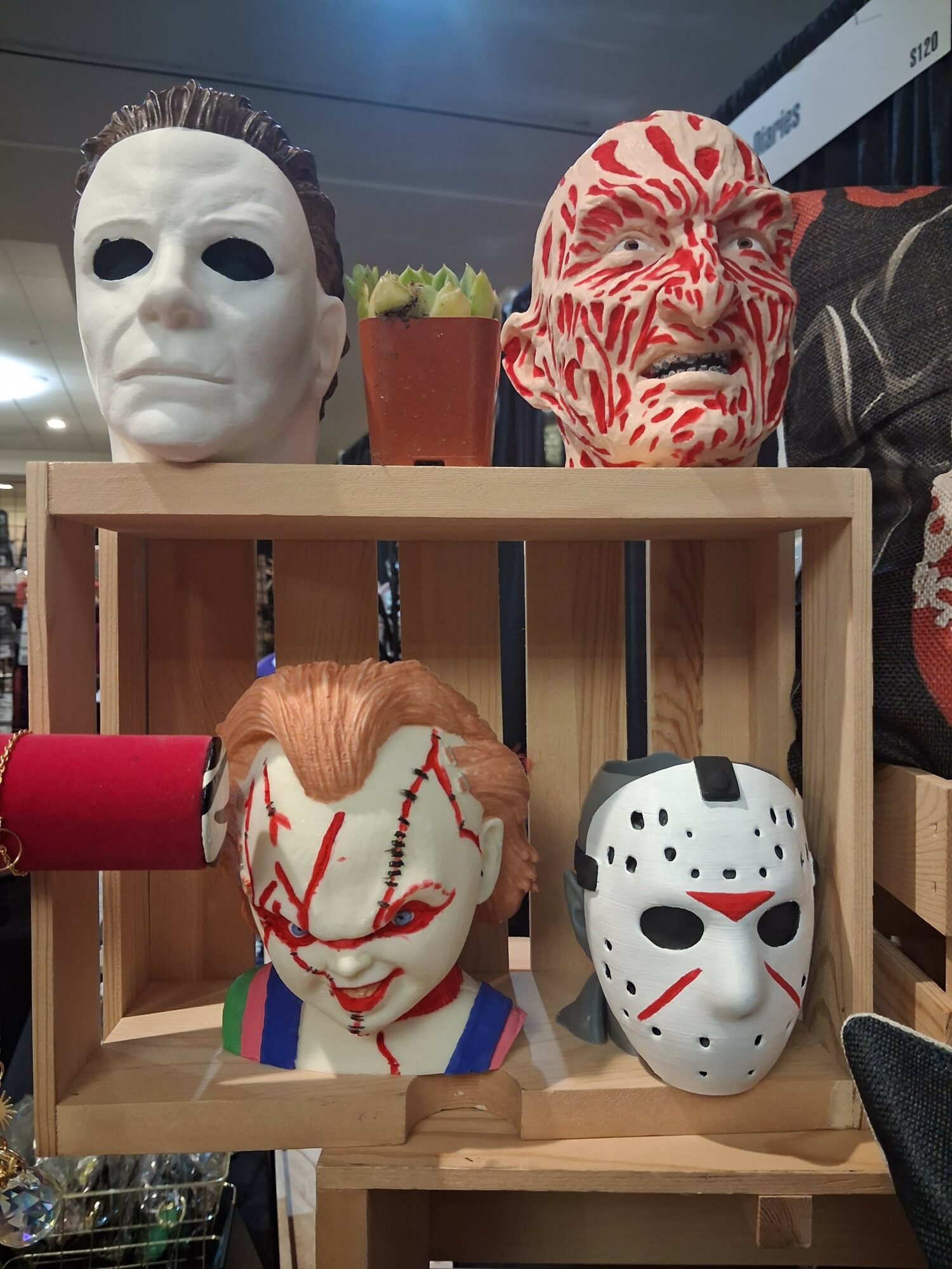 Three masks and a plush toy inside a wooden box, with a person wearing a patterned shirt nearby.