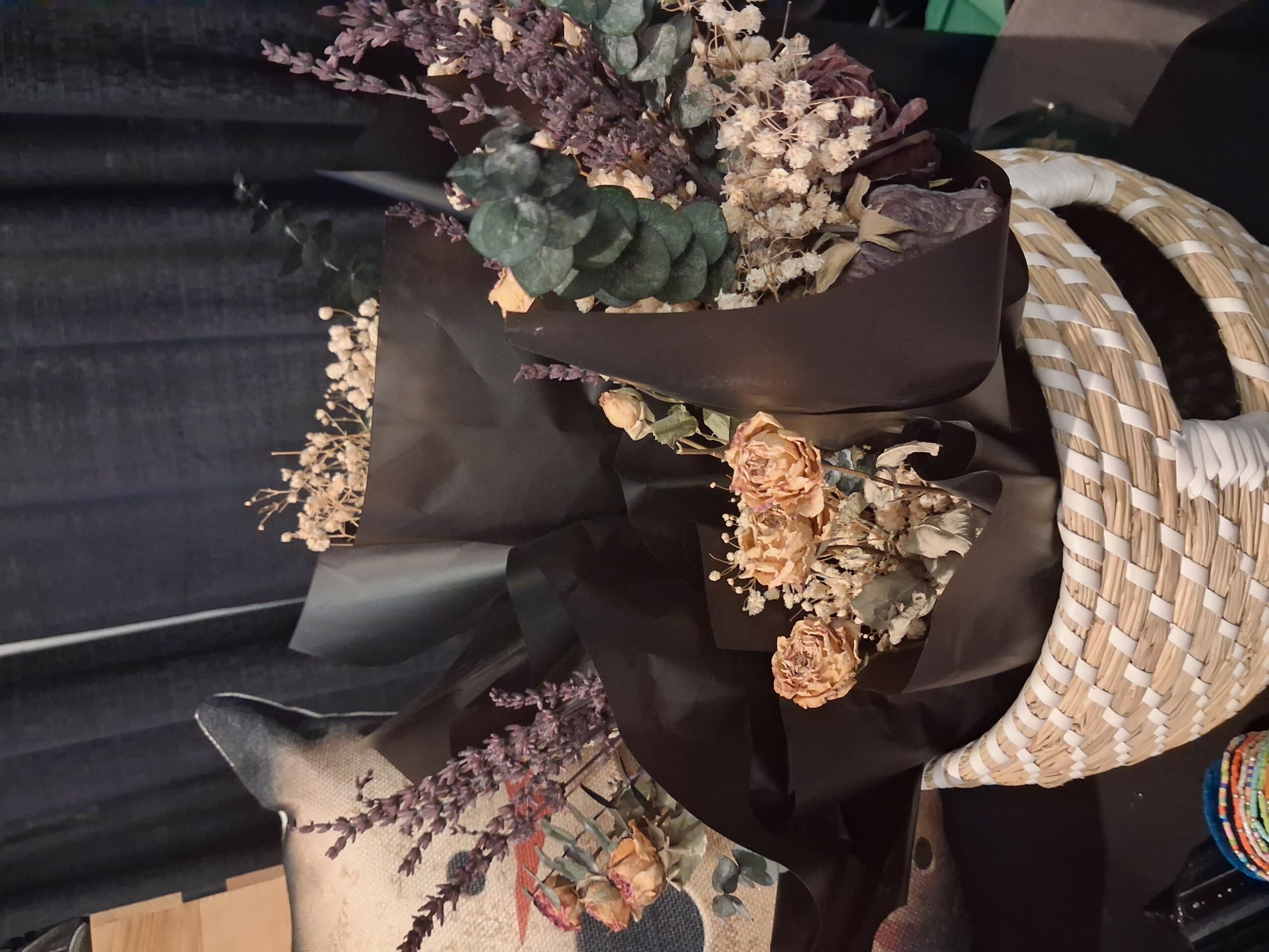 Bouquet of dried flowers wrapped in black paper, placed in a woven basket, with a dark background.