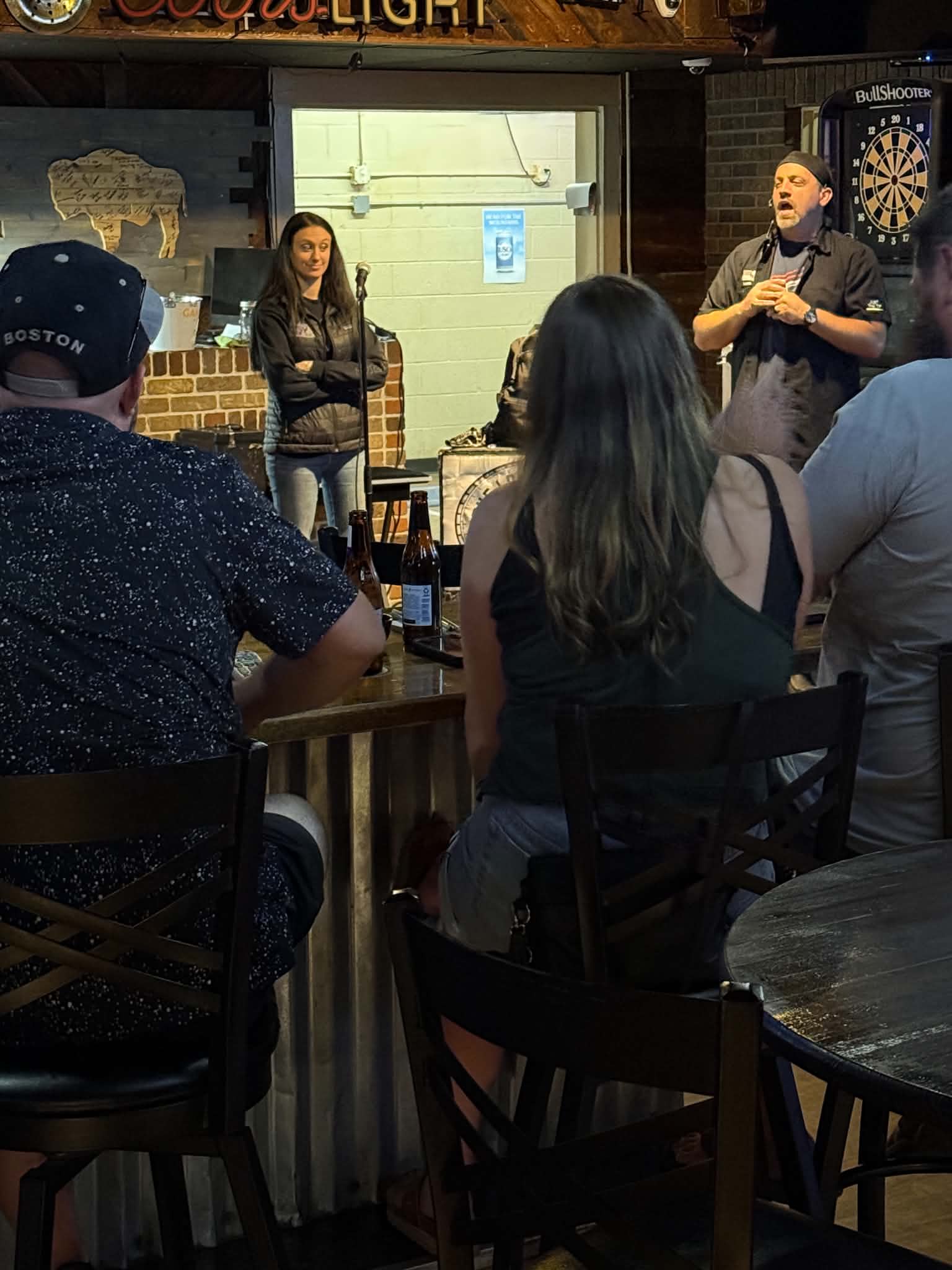 People seated at tables listening to a speaker in a bar or pub setting, with a dartboard on the wall.