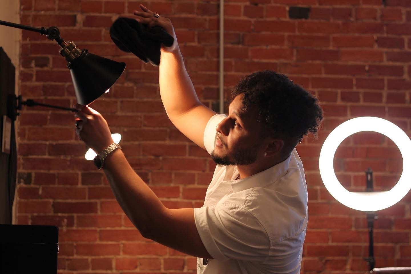 Young man adjusting a studio light in front of a brick wall, illuminated ring light visible in background.
