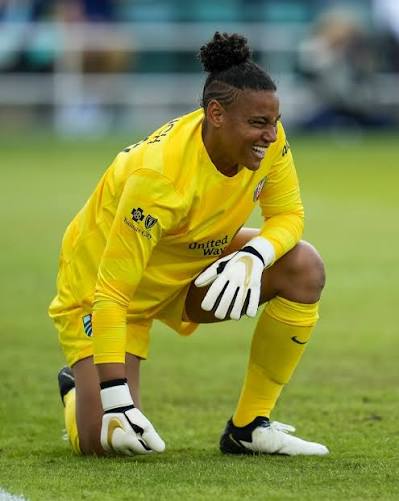 Female soccer player in yellow uniform crouching on grass field, smiling, wearing gloves and cleats.