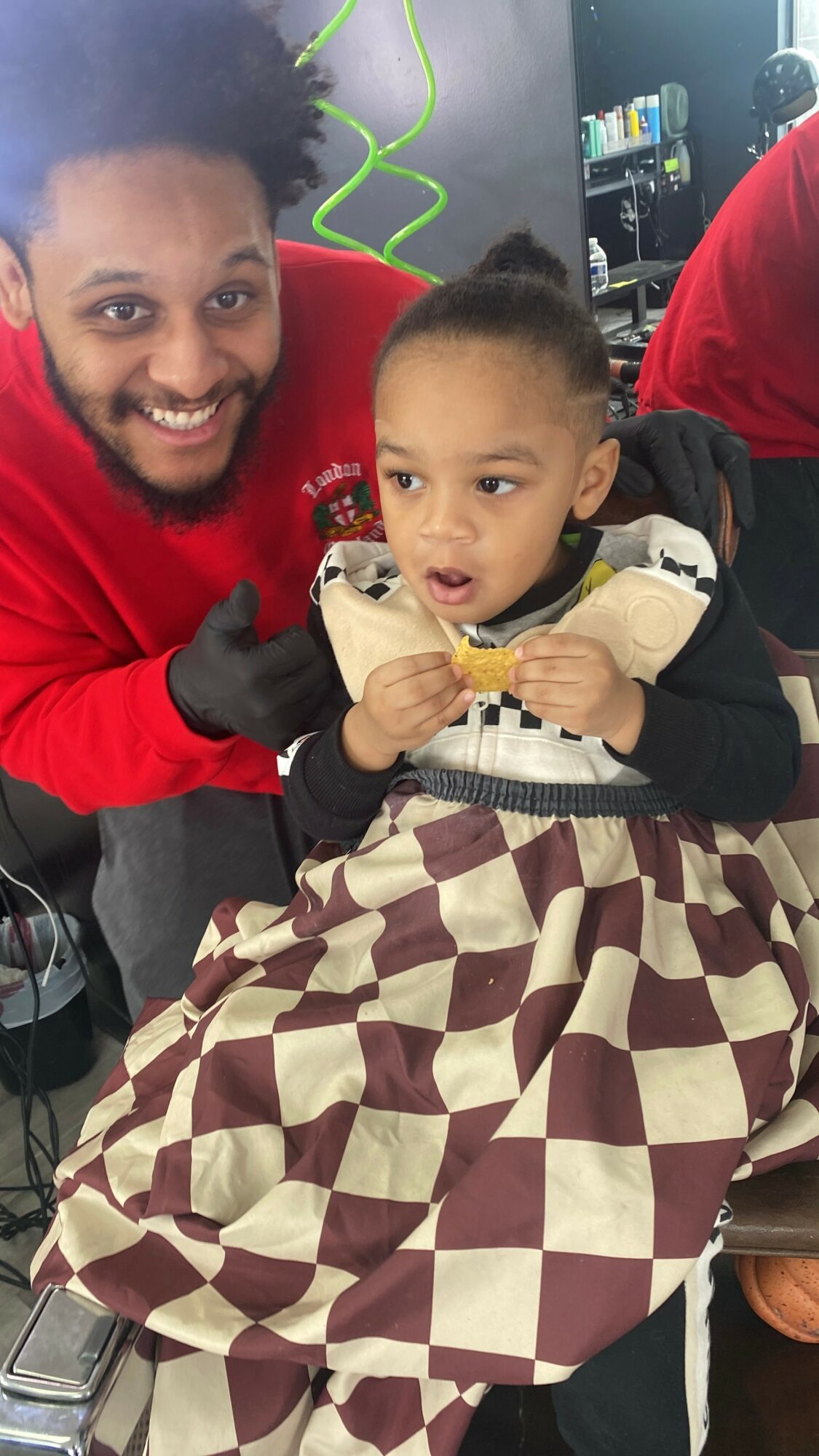 Smiling man with curly hair and a child sitting on a chair, holding a snack, in an indoor setting.