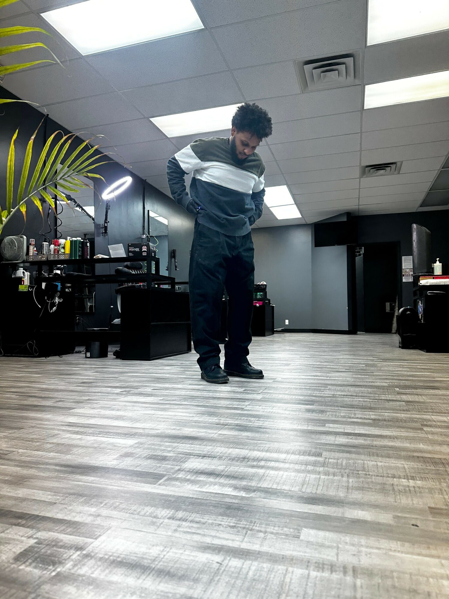 Person standing in a room with a wooden floor, facing a wall with ceiling lights, with various objects and furniture around.