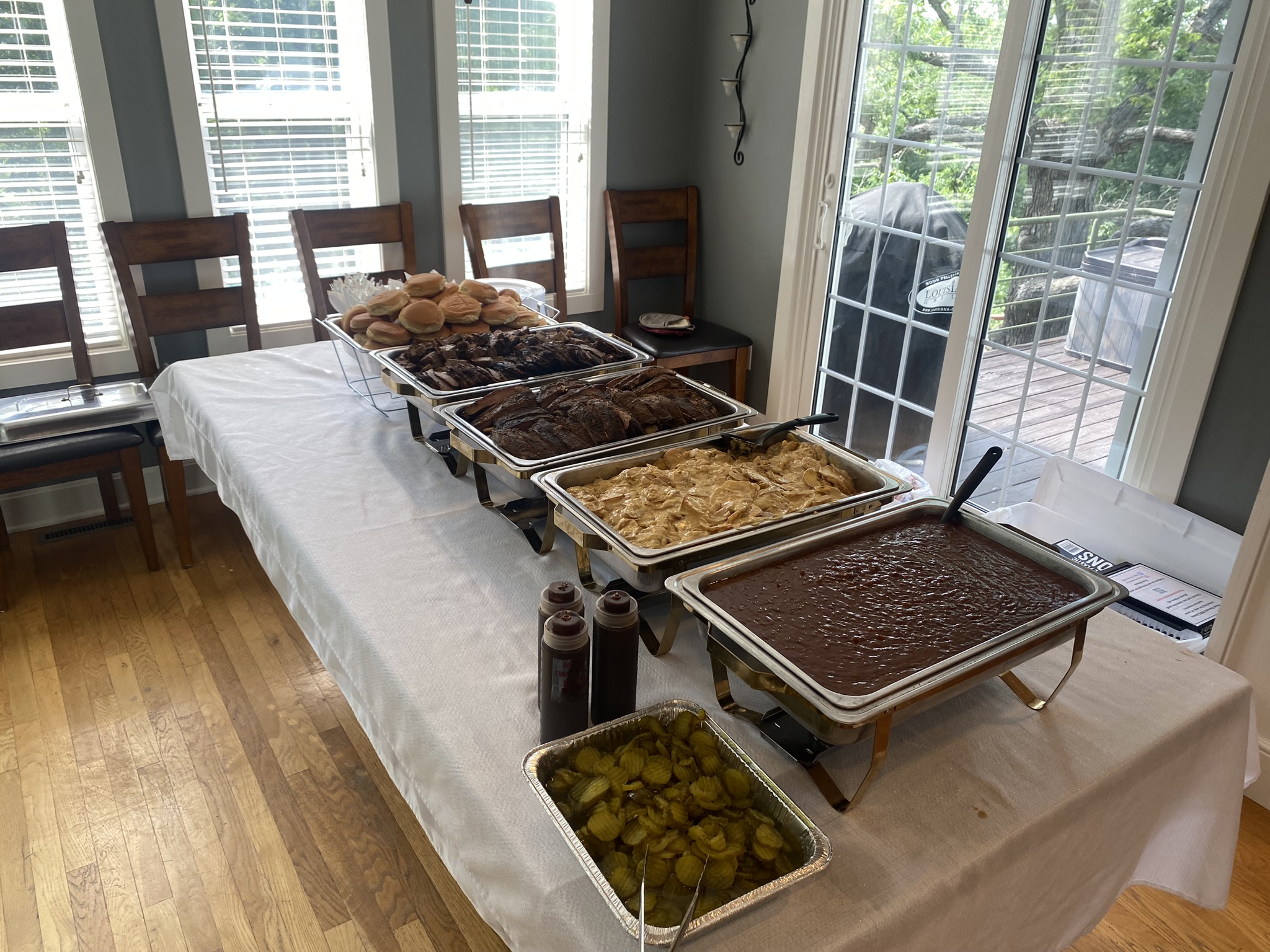Buffet table with various dishes including baked goods, casseroles, and drinks, set in a dining area with chairs and windows.
