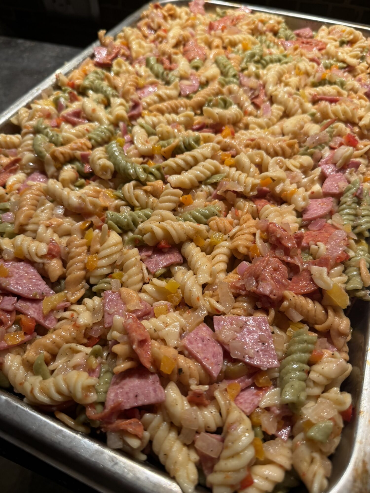 Close-up of a large tray filled with colorful pasta salad containing rotini pasta, ham, and vegetables.