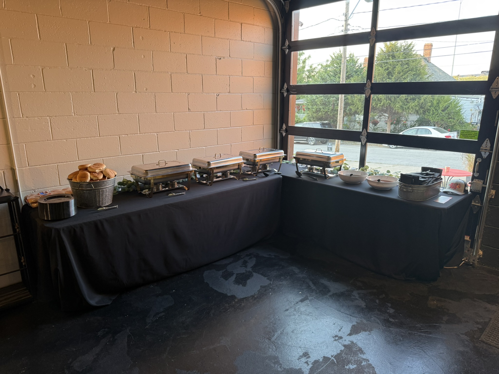 Long table with food and dishes near a large window, brick wall in background, dark floor, natural light.