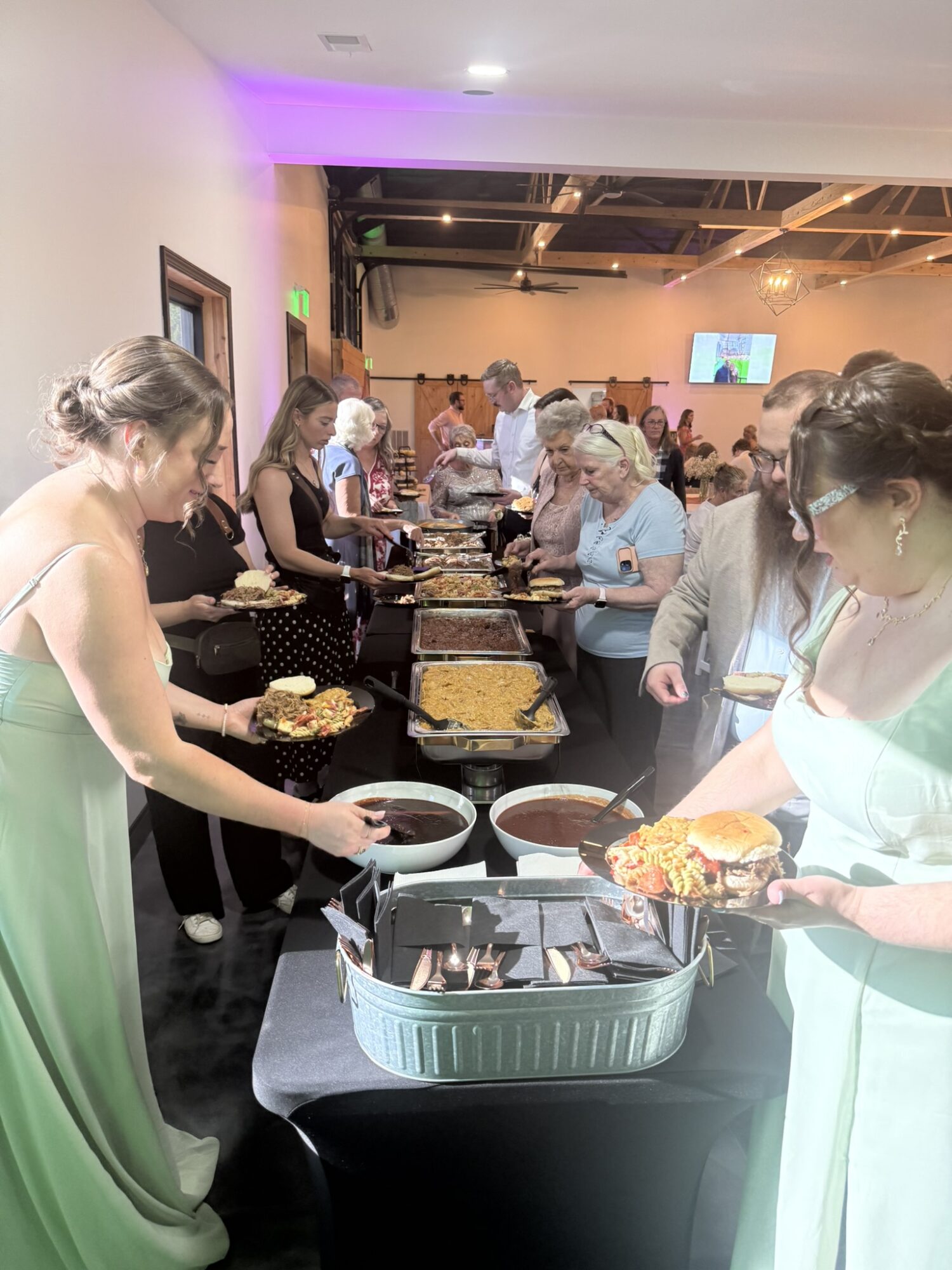 People line up at a buffet table with food trays in a well-lit indoor setting, some serving themselves. Group of women in formal attire.