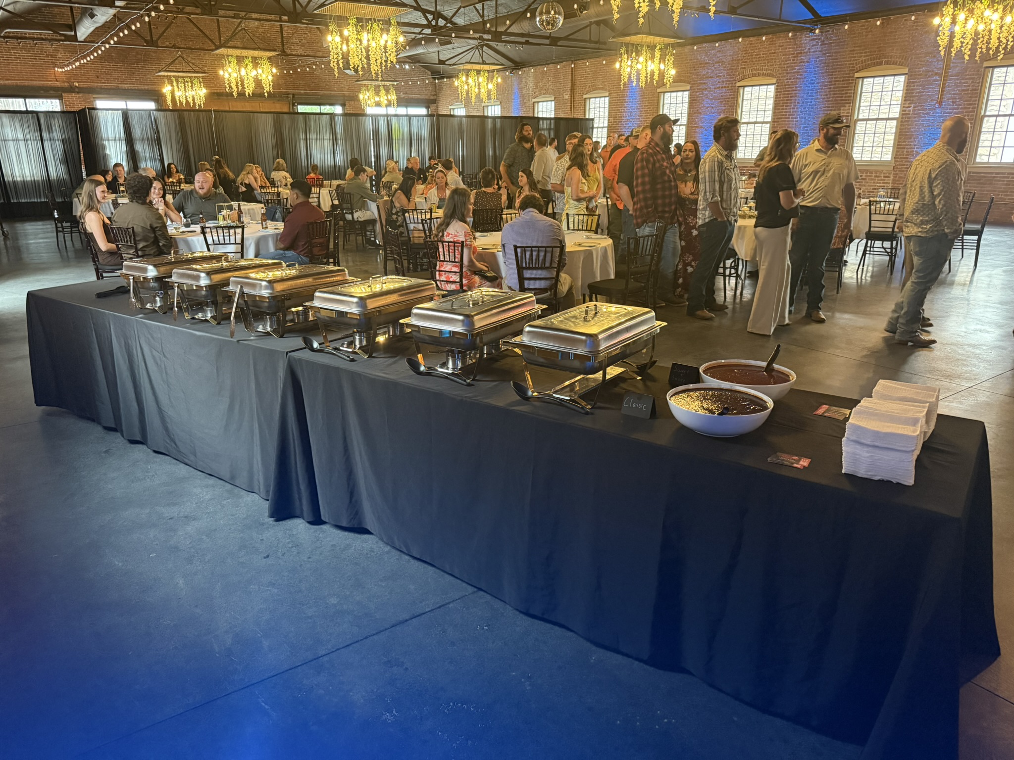 Buffet table with chafing dishes, bowls, and stacks of plates in a large banquet hall with people dining and socializing.