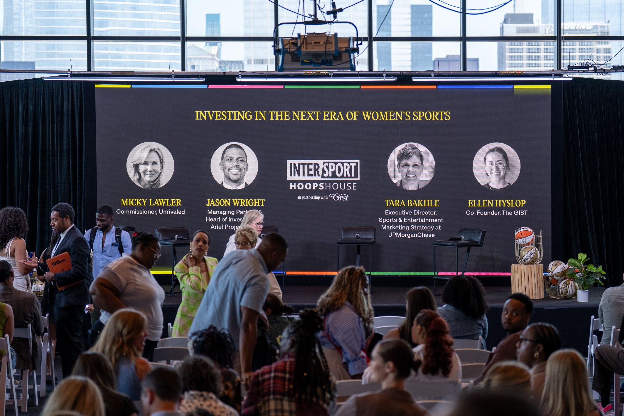 Audience seated in front of a stage with a large screen displaying images and names of women sports figures, and the InterSport Hoops House logo.