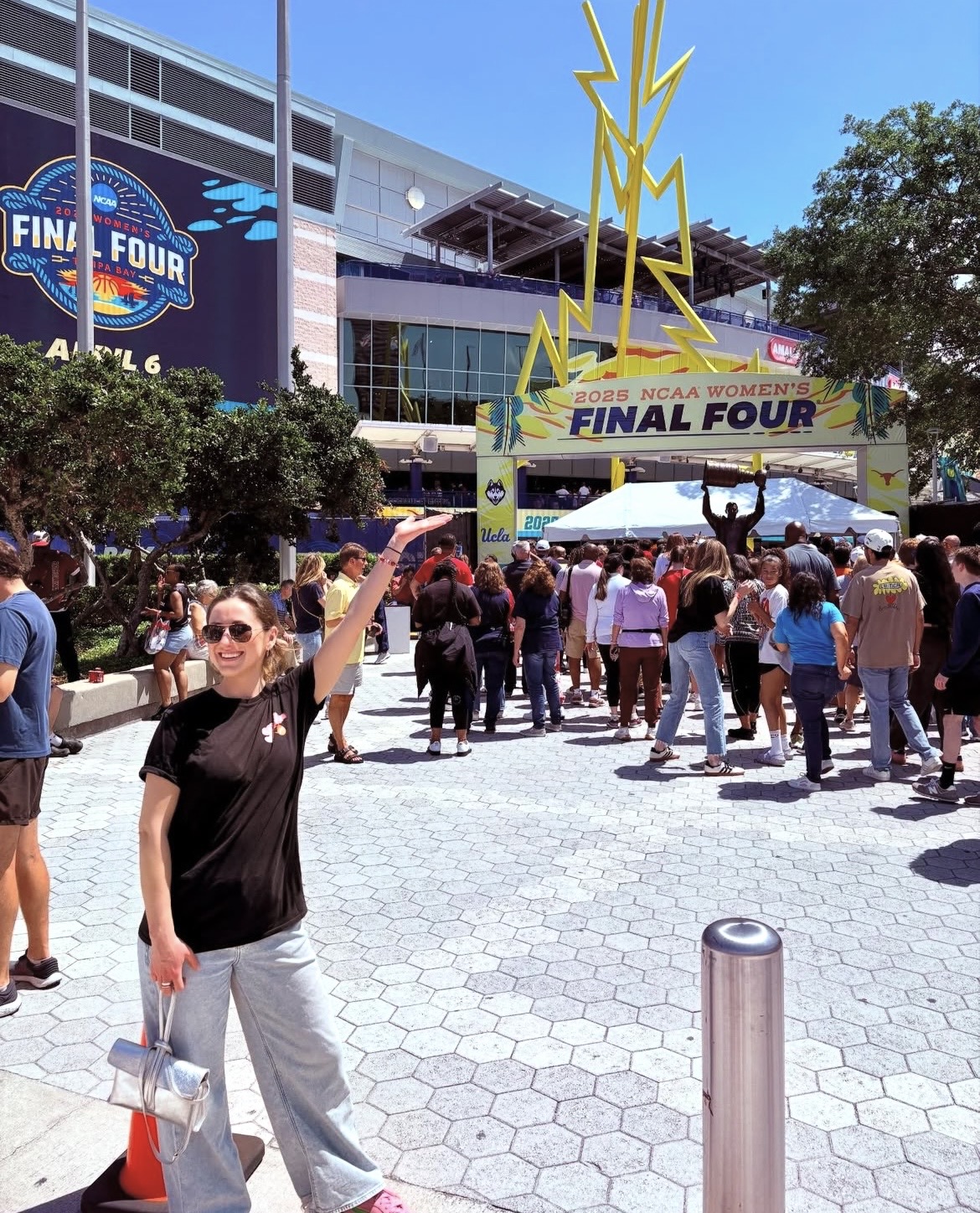 Person in black shirt and gray pants holding a bag, standing in crowd outside stadium for NCAA Women's Final Four, sunny day.