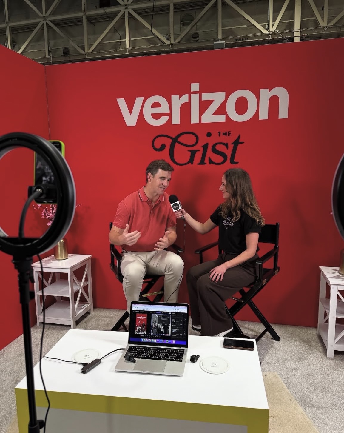 Two people sit and talk during an interview with a red Verizon Gist backdrop, a laptop, and lighting equipment present.