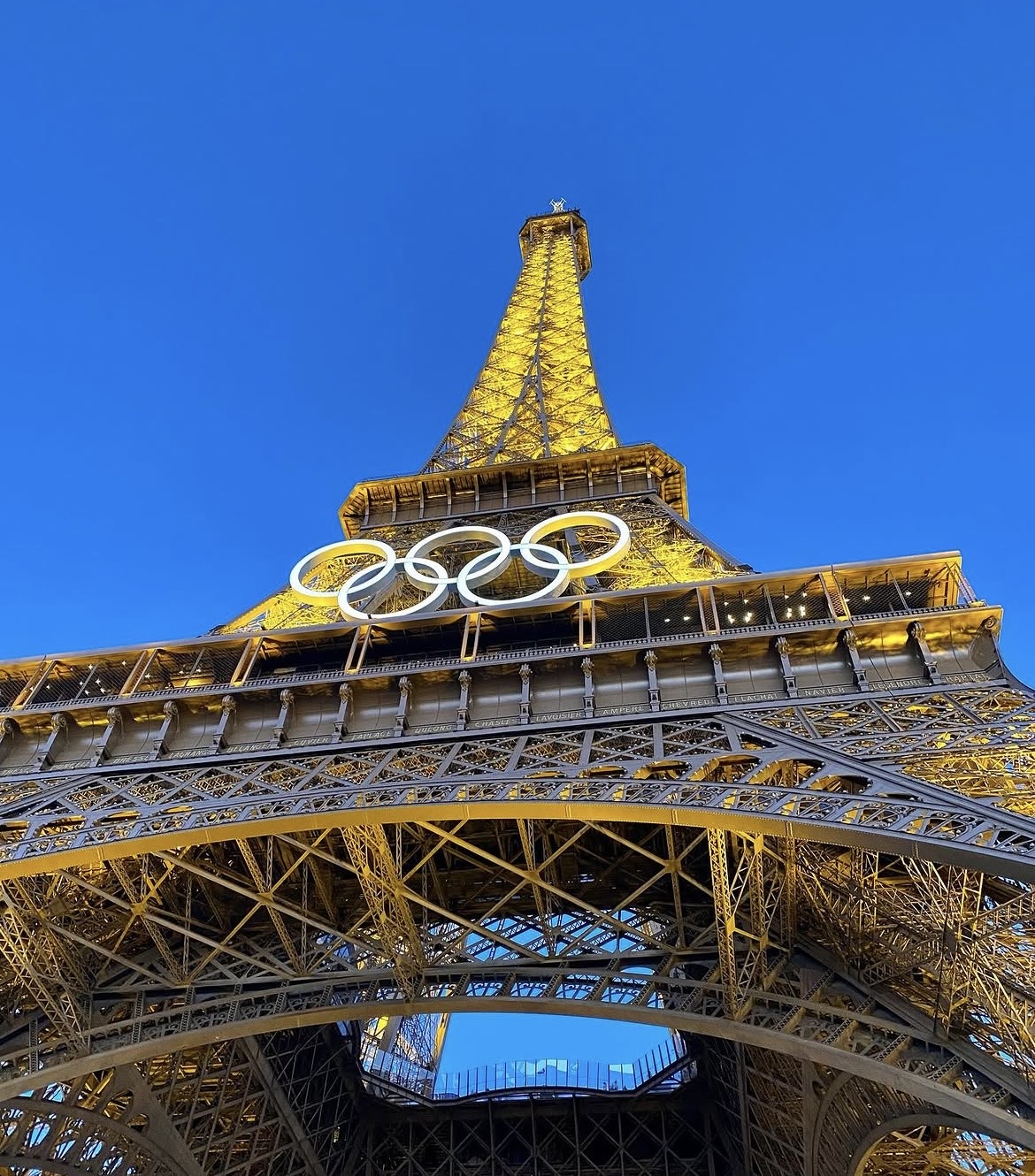 Eiffel Tower illuminated at night with Olympic rings hanging below, viewed from below against a clear blue sky.