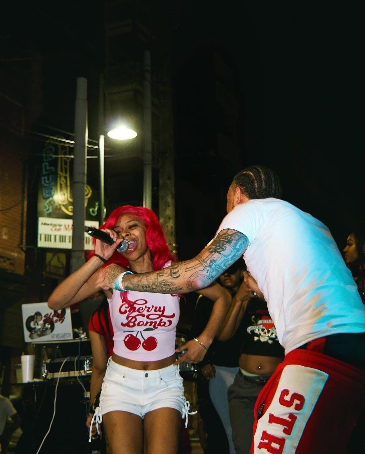 Two people on stage, one with red hair and a microphone, the other with tattoos, in a nighttime outdoor setting.