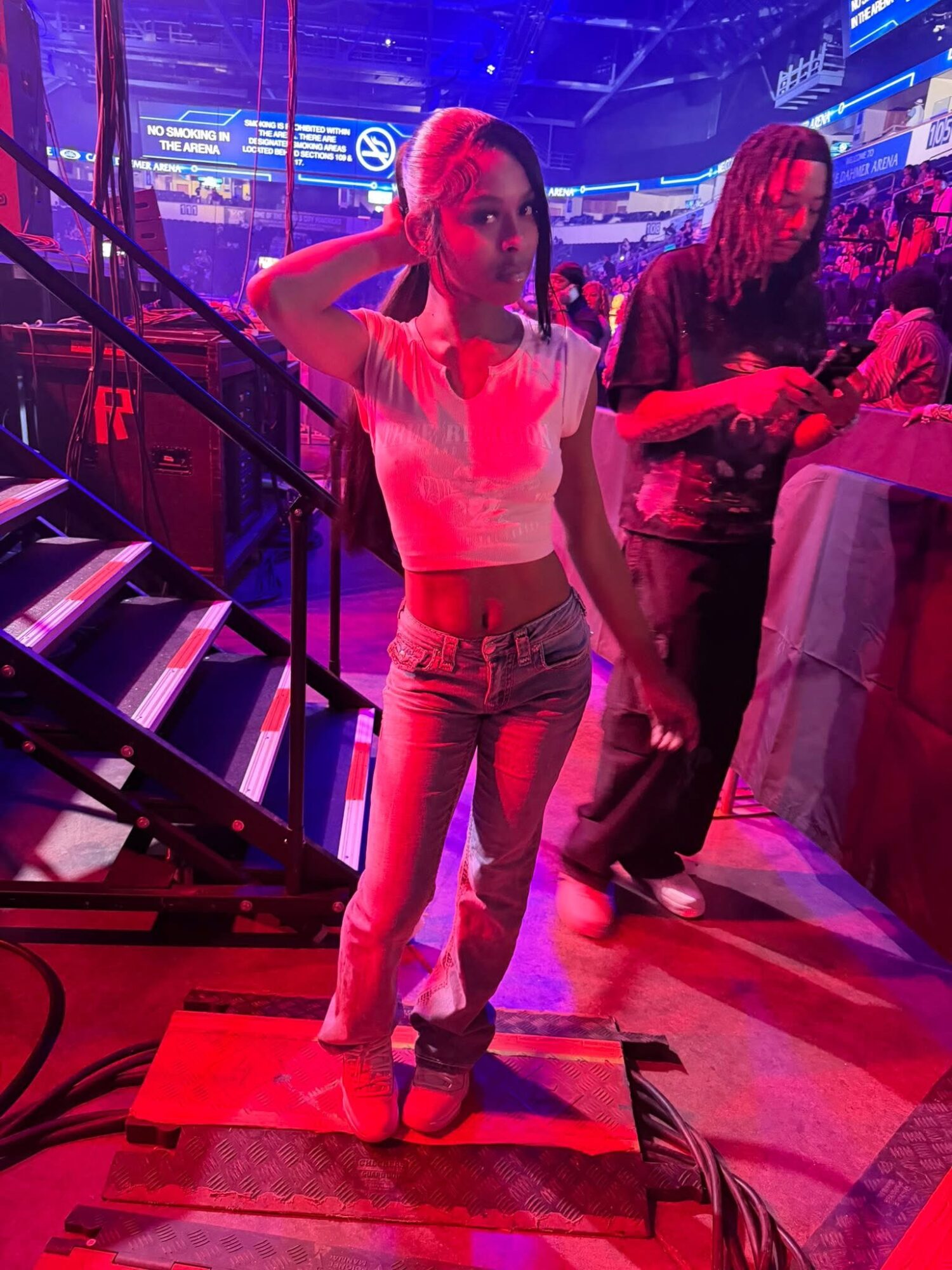 Young woman standing on a stage with colorful lights, wearing a white top and jeans, in a lively indoor setting.