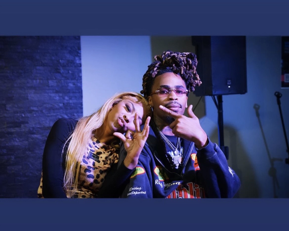 Two people sitting together, one with long blonde hair and the other with curly hair and glasses, in a room with a dark wall and speakers.