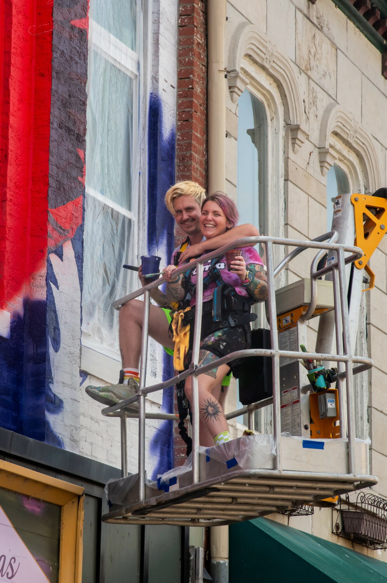 Two people smiling and hugging on a raised platform in front of a building with graffiti and ornate architecture.