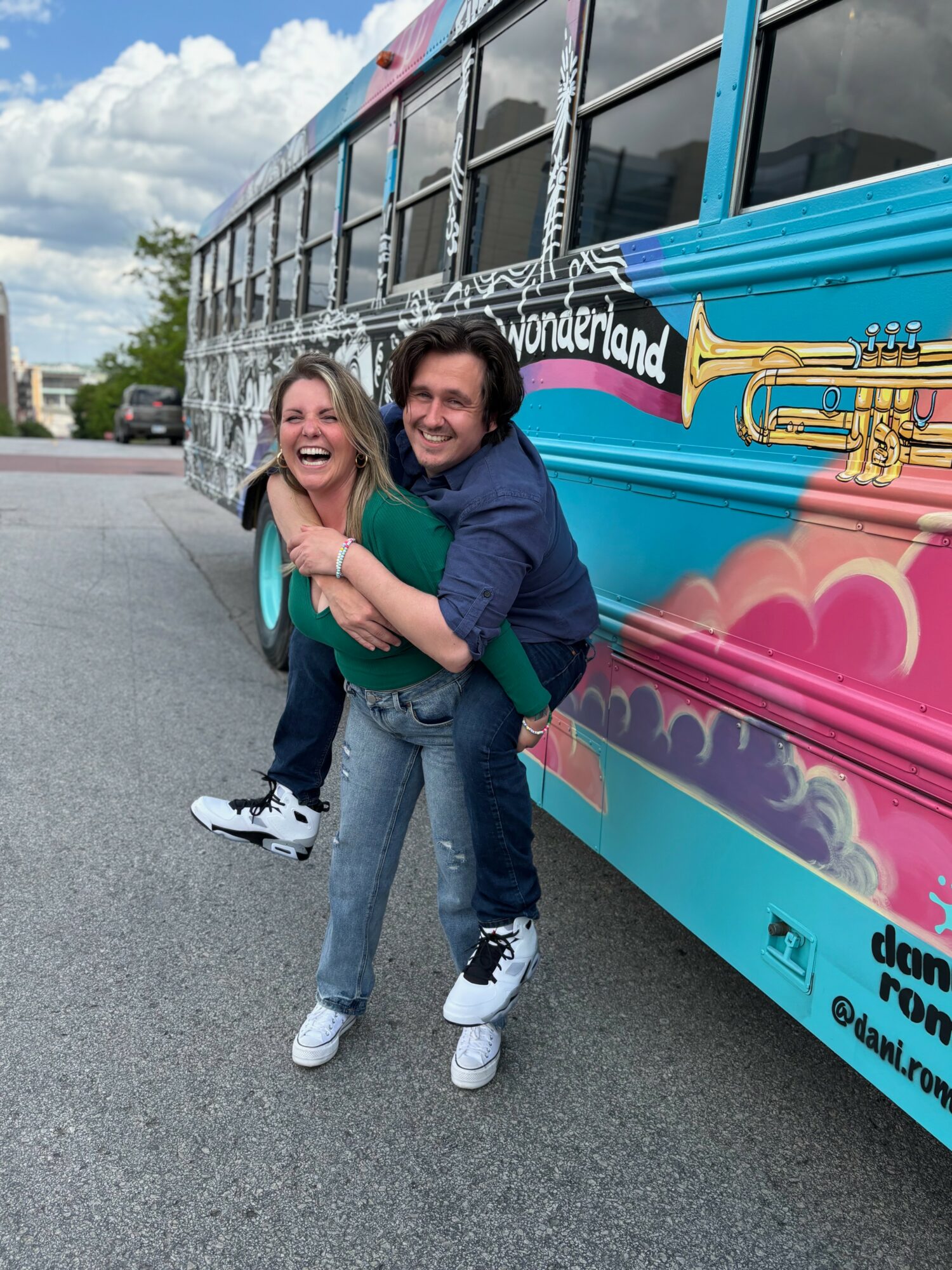Two people smiling and hugging in front of a colorful bus with a Wonderland sign, cloudy sky in background.