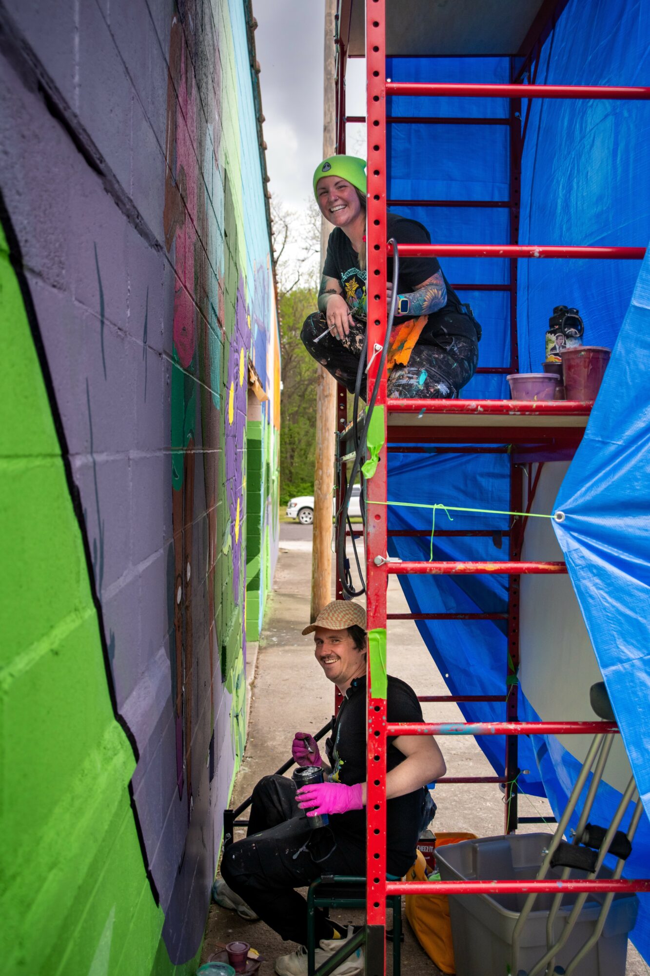 Two children climbing a colorful outdoor structure, one smiling and wearing a green hat, the other sitting with a hat, against a mural background.