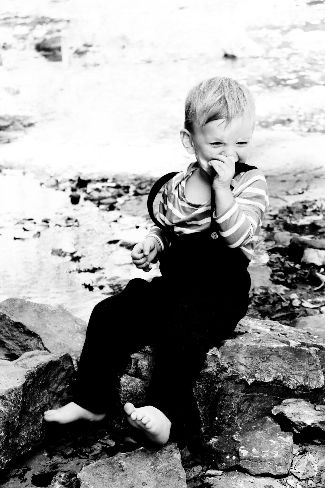 Child sitting on rocks near water, smiling and holding hand to mouth, wearing striped shirt and dark pants.