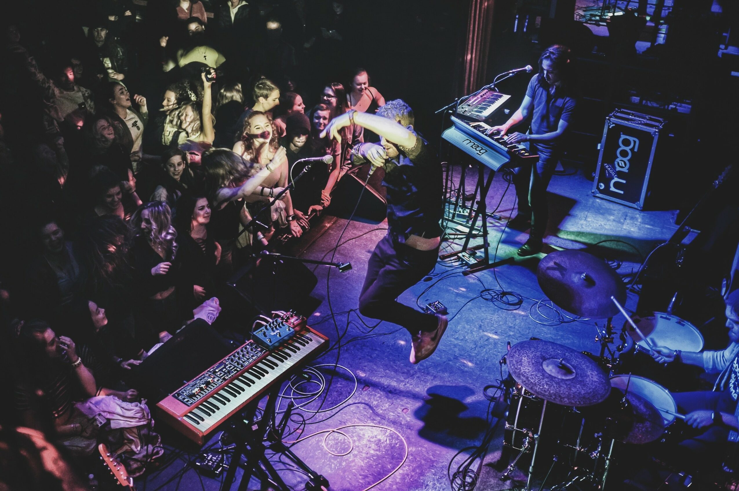 Musicians perform on stage with a keyboard, drums, and a singer, while an audience watches attentively in a dimly lit venue.