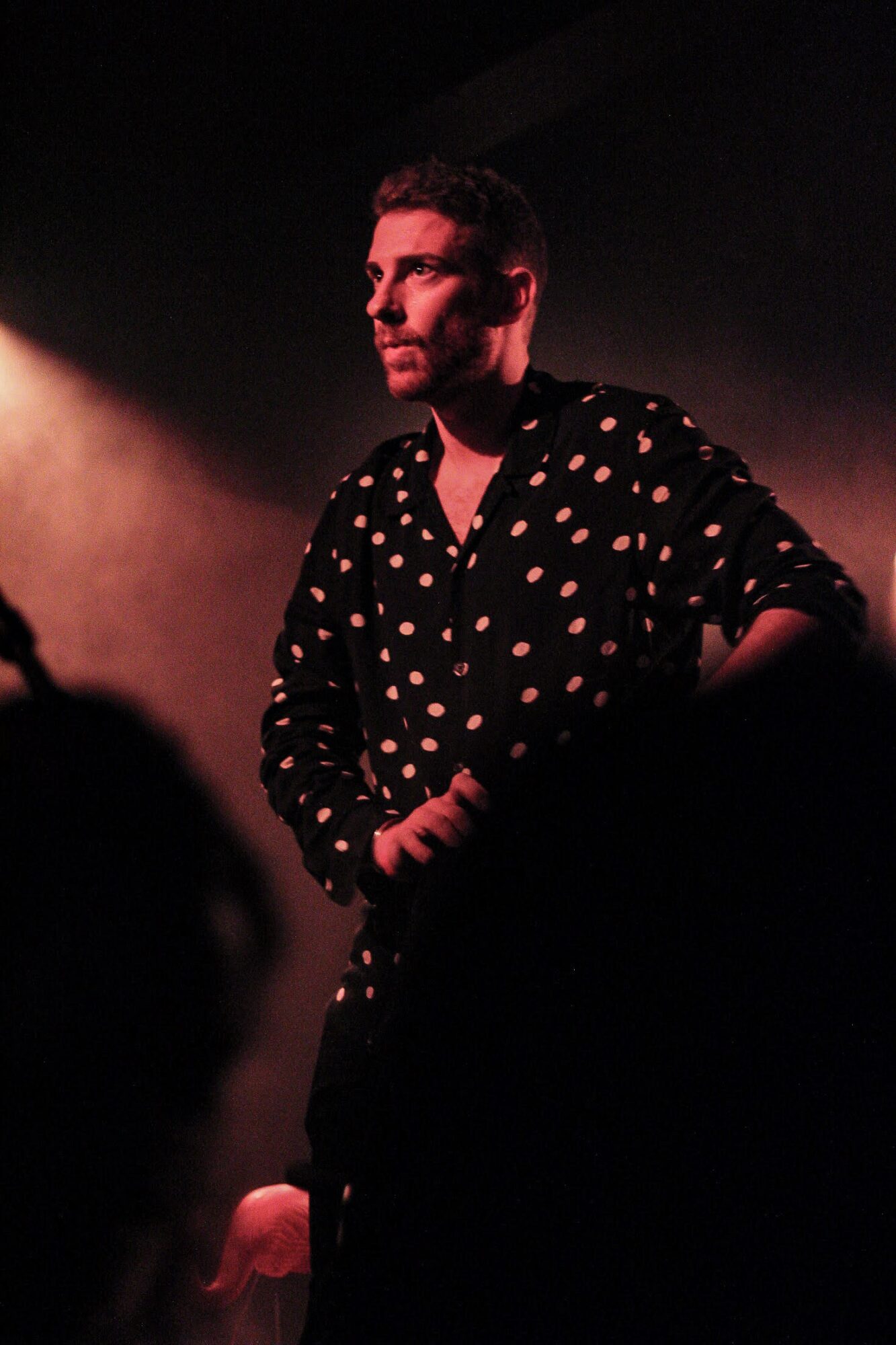 Person standing on stage with dark background, wearing a black shirt with white polka dots, illuminated by stage lighting.