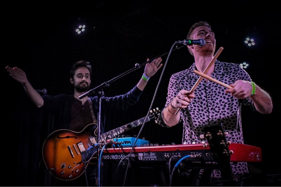Two musicians perform on stage, one singing into a microphone and playing drums, the other with a guitar and keyboard.