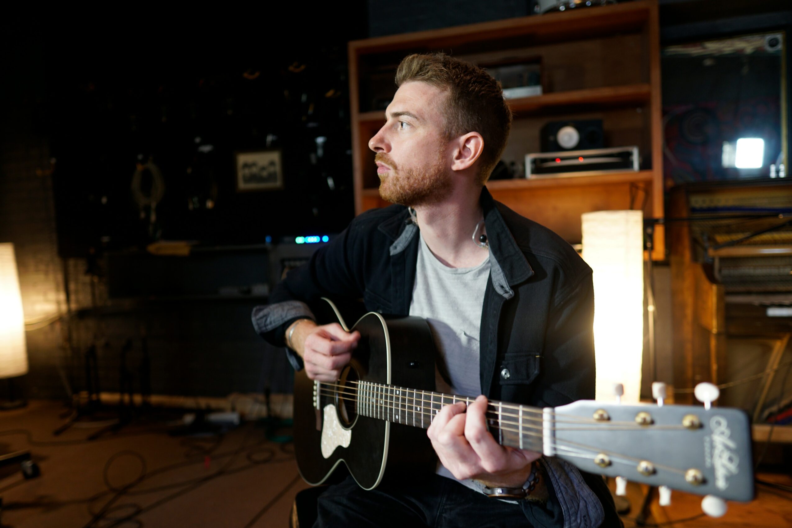 Man with a beard playing an acoustic guitar in a dimly lit room with shelves and a TV behind him.