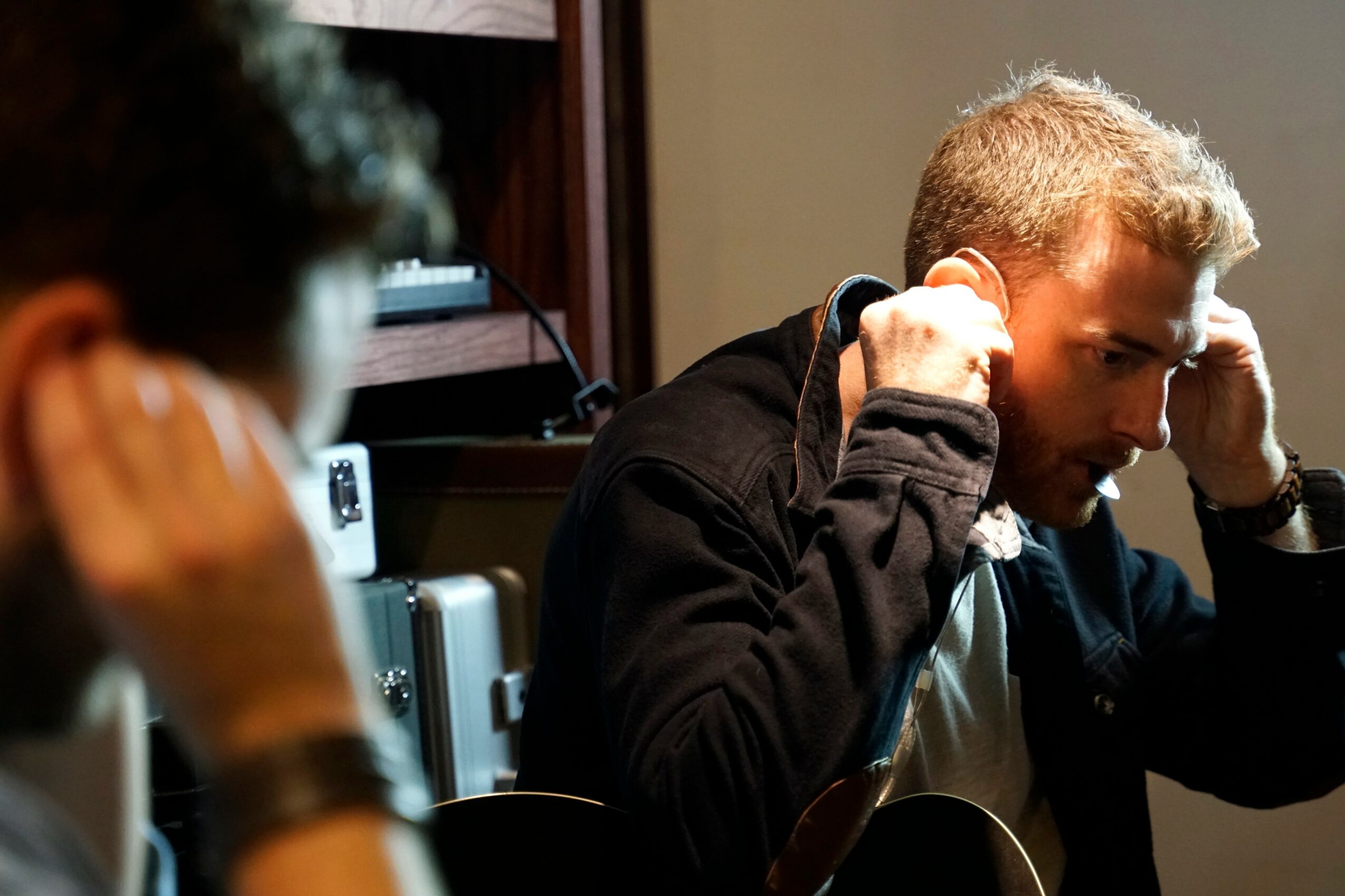 Two people are engaged in a conversation, one with hands on head, in a room with shelves and electronic equipment.
