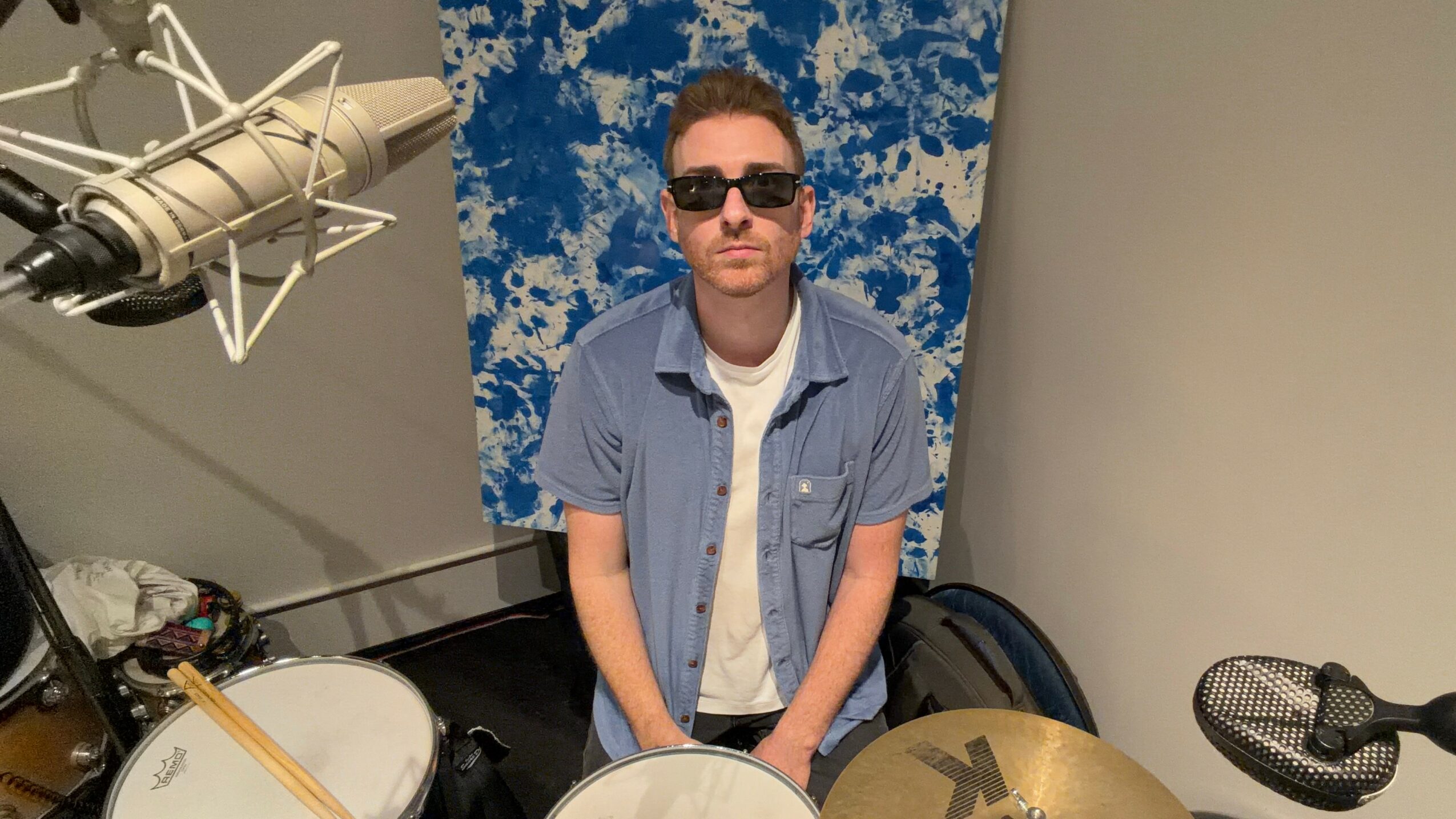 Person wearing sunglasses and a denim shirt, sitting in a music studio with drums and microphones.