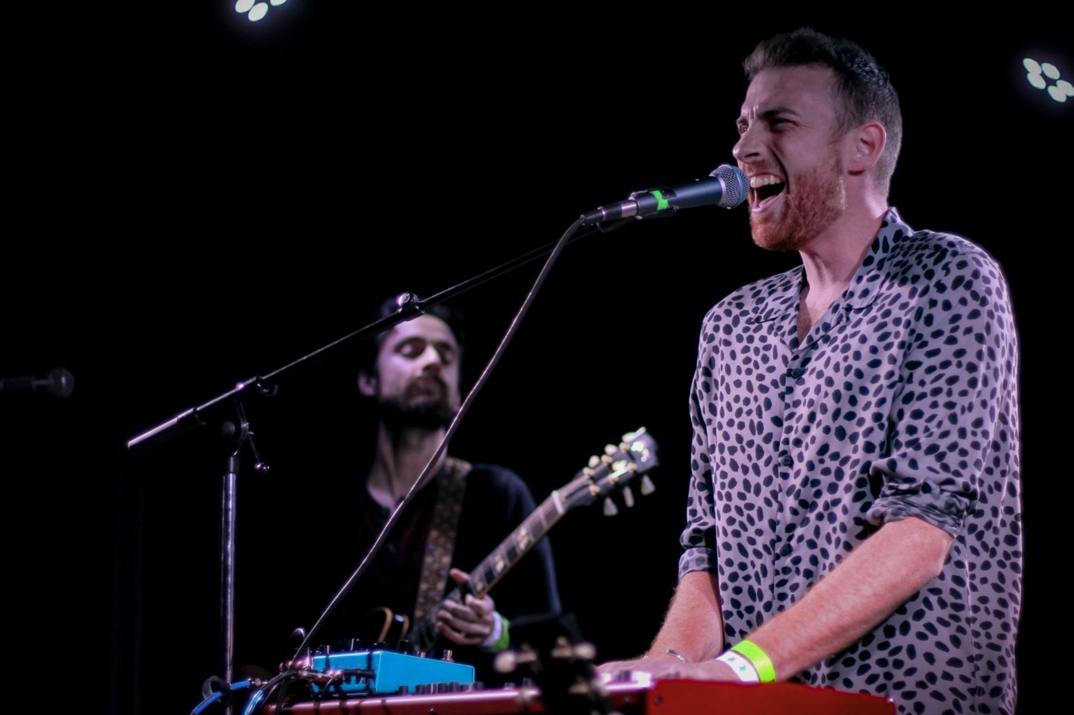 Two men perform on stage, one singing into a microphone and the other playing guitar, with stage lights overhead.