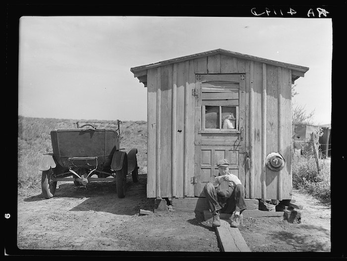 Person sitting outside a small wooden shed with a window, an old car parked nearby, and a dog on the ground.