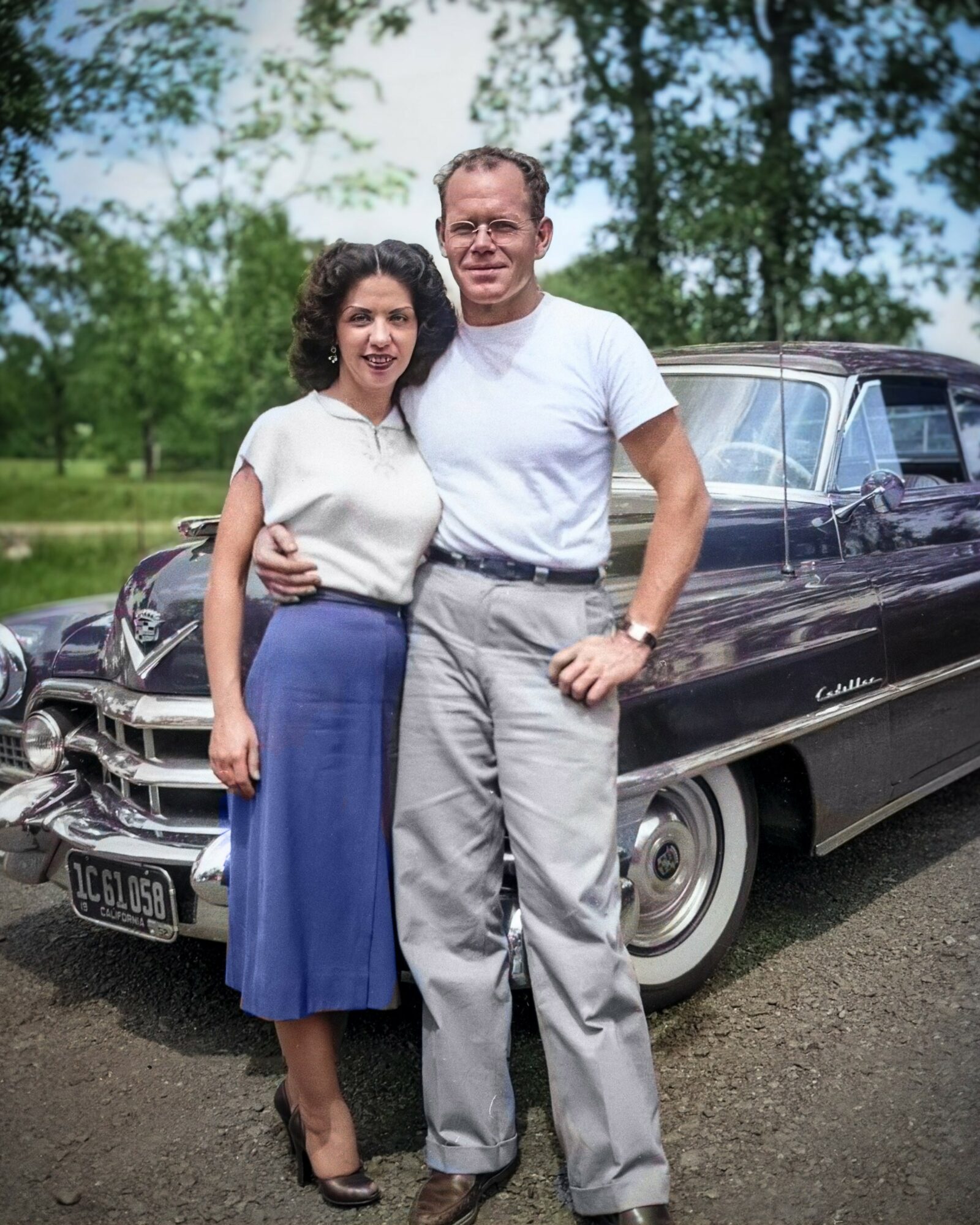 A man and woman stand outdoors in front of a vintage car, smiling, with trees and blue sky in the background.