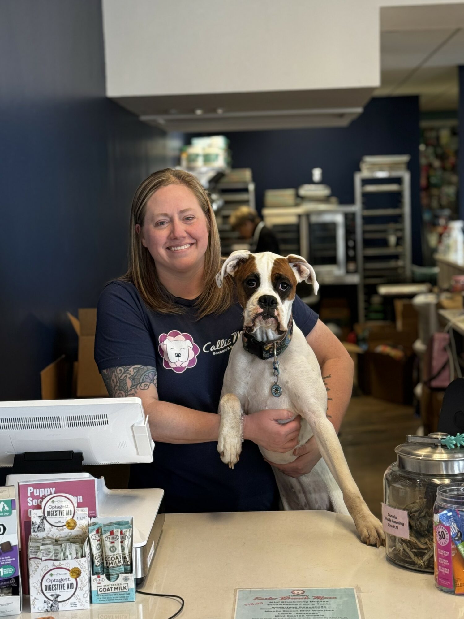 Woman smiling holding a dog at a counter in a store, with shelves and products in the background.