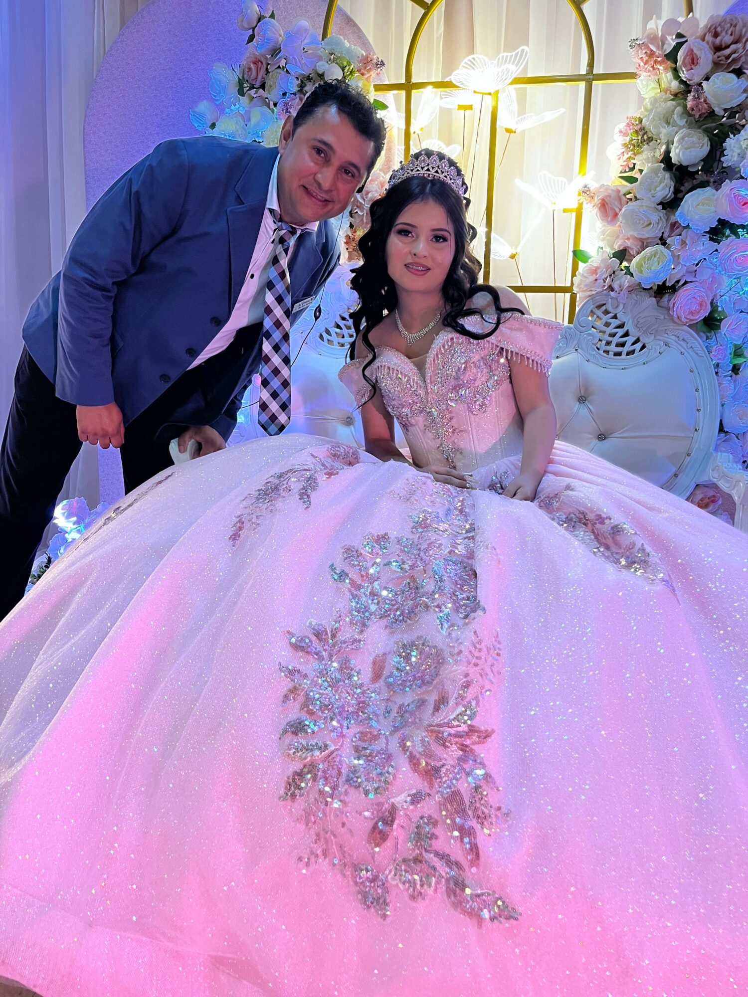 A couple dressed in formal attire, standing close together at a decorated event with floral arrangements and lighting.