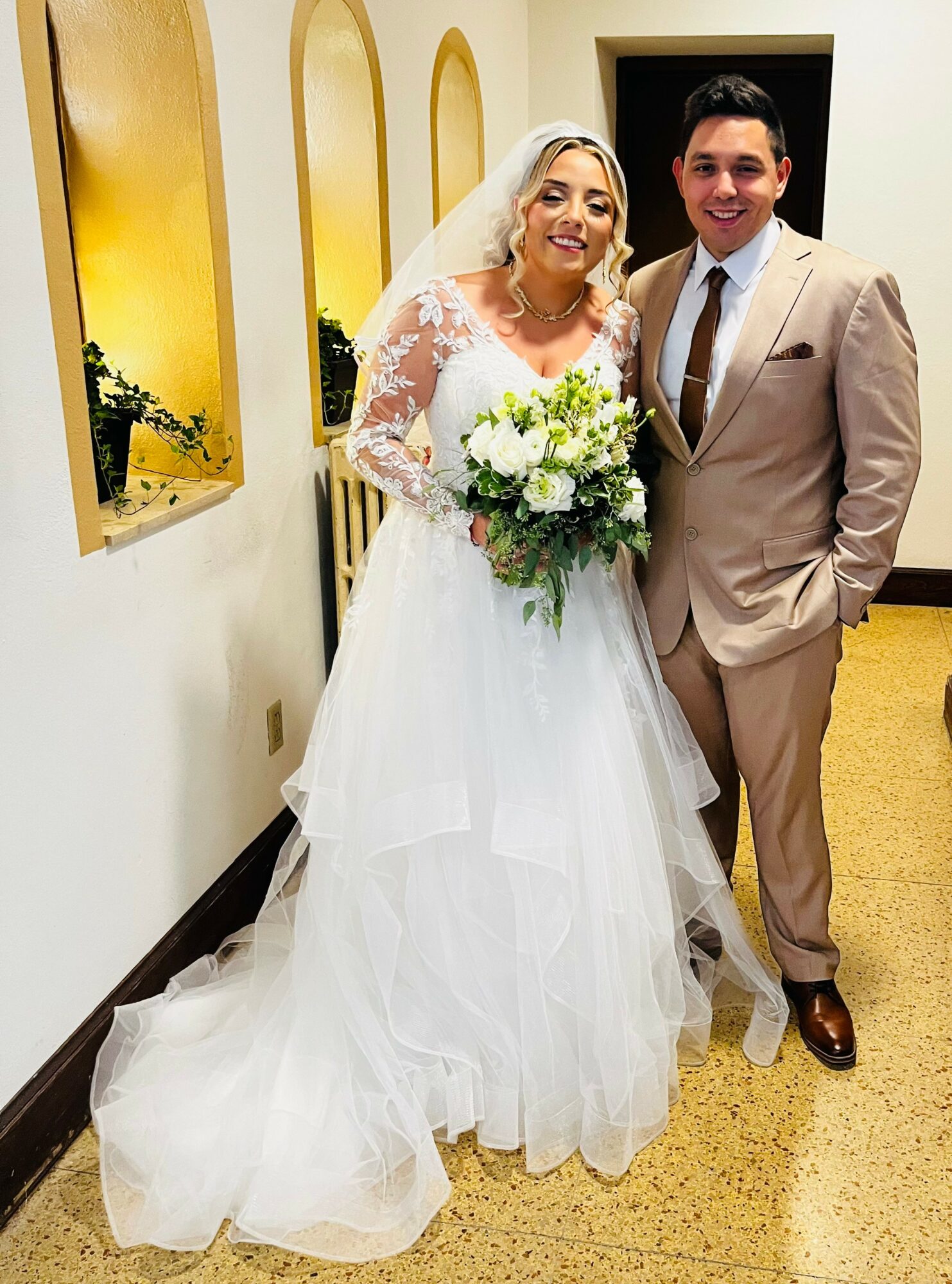 Bride in white wedding dress holding bouquet, groom in beige suit standing together indoors, smiling.